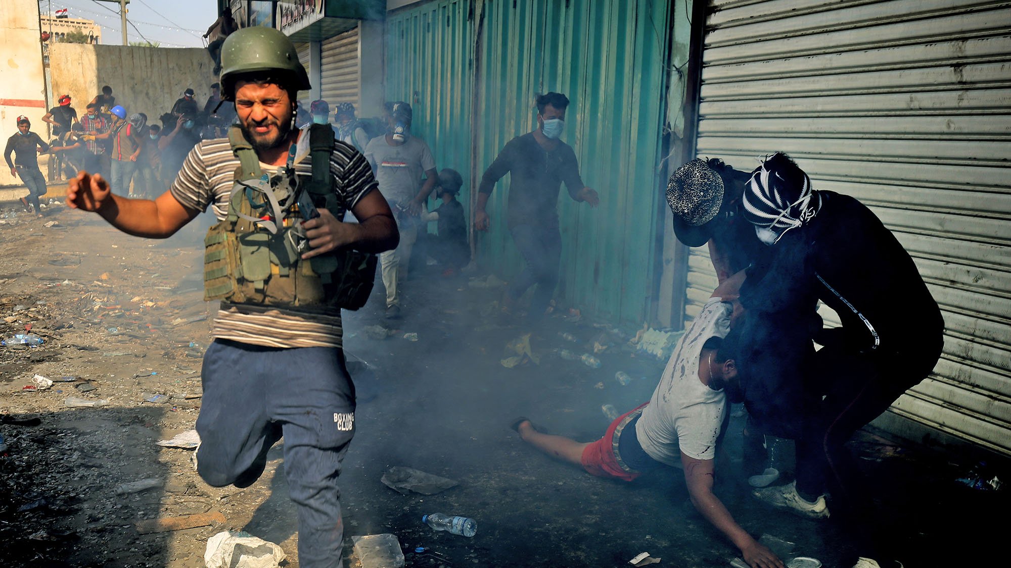 تظهر الصورة مشهدًا من الفوضى، حيث يركض رجل يرتدي خوذة ودروعًا واقية وسط دخان كثيف في منطقة يبدو أنها تعرضت لأعمال شغب أو احتكاك. في الخلفية، يظهر عدد من الأشخاص، يبدو أن بعضهم يتفاعل بشكل عاطفي مع الوضع. الأرض مليئة بالقمامة، مما يعكس حالة من الاضطراب والانزعاج. الجو العام يوحي بالتوتر والخطر.