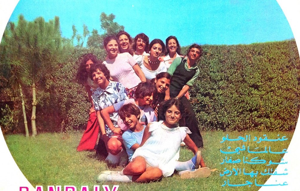 L'image représente une couverture de disque avec un groupe de personnes posant ensemble à l'extérieur, probablement dans un jardin. Ils semblent s'amuser et sourire, créant une ambiance joyeuse. Le nom du groupe, "BANDALY", et le titre "The Merry Family" sont présentés en bas, avec des inscriptions en arabe. En arrière-plan, on peut voir des arbres et une haie. L'ensemble évoque une atmosphère festive et conviviale.