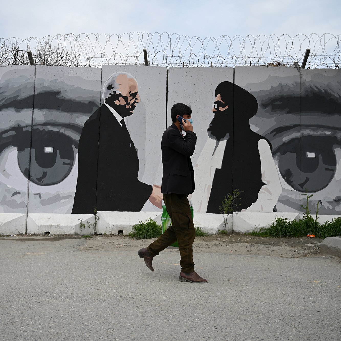 L'image montre un homme marchant sur une route, portant un costume sombre, tandis qu'il téléphone. En arrière-plan, on aperçoit un mur décoré d'une fresque qui représente des visages stylisés, probablement en noir et blanc. L'œuvre met en avant des yeux perçants et deux silhouettes, ce qui semble évoquer des figures significatives. La scène dégage une atmosphère de tension et de réflexion, juxtaposition entre le monde urbain et l'art mural. Des barbelés sur le mur ajoutent une dimension de restriction ou de sécurité.