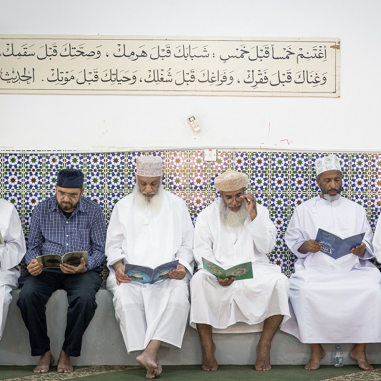 L'image montre un groupe d'hommes assis en ligne, lisant des livres, probablement des copies du Coran. Ils portent des vêtements traditionnels, tels que des dishdashas et des couvre-chefs. En arrière-plan, on peut voir un mur décoré de carrelage coloré, sur lequel est inscrite une citation ou un texte. L'atmosphère semble calme et sereine, suggérant un moment de prière ou de méditation.