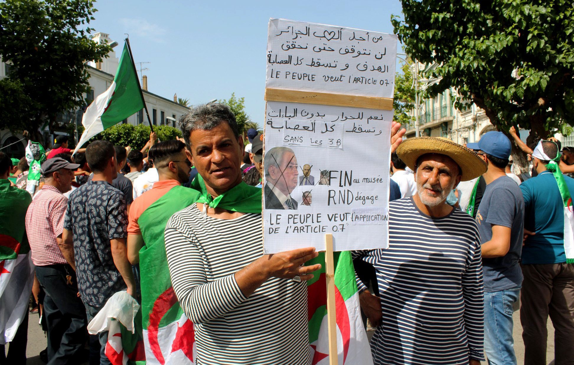 تظهر في الصورة مجموعة من الأشخاص في تجمع أو مظاهرة. يحمل بعضهم لافتات تتضمن شعارات سياسية تتعلق بالمطالب الشعبية في الجزائر. يرتدي المشاركون ملابس بألوان متنوعة، ويبدو أن هناك استخداماً للعلم الجزائري. المشهد يعكس روح الحماس والتعبير عن المطالب الجماهيرية.