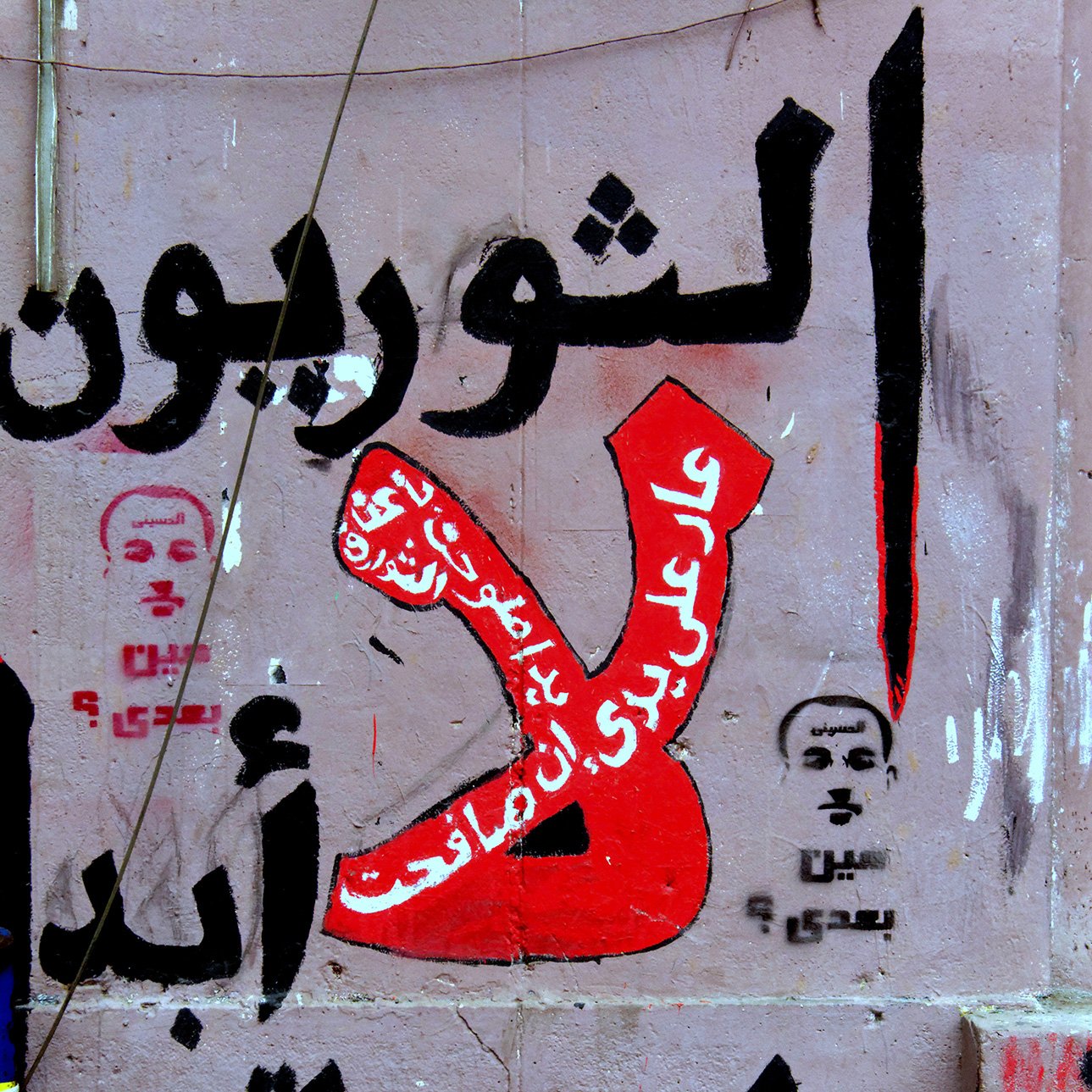 L'image montre un mur recouvert de graffitis, avec des inscriptions en arabe. On peut y voir le mot "Non" stylisé en rouge, entouré de phrases opposées à certaines idées ou actions. Des visages ou portraits, probablement des figures publiques, sont également esquissés. L'ensemble de la composition véhicule un message fort et engagé, typique de la communication visuelle dans des contextes de protestation sociale ou politique.