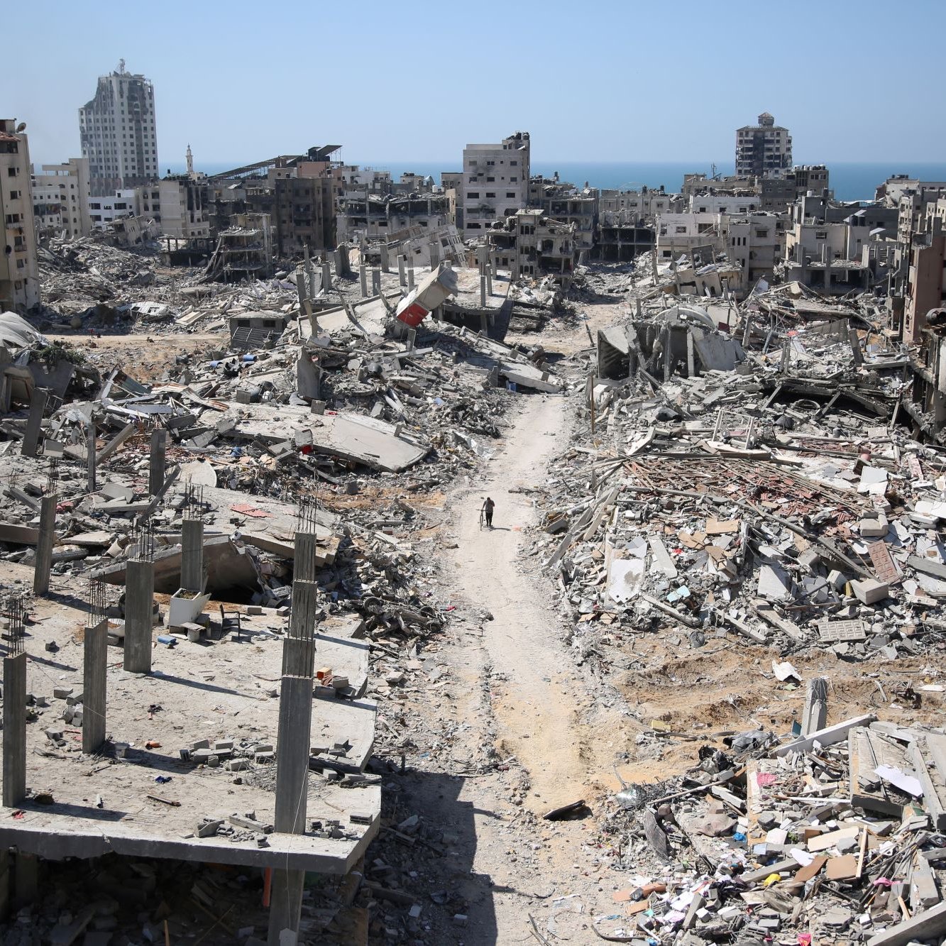 L'immagine mostra un paesaggio di devastazione, con edifici in rovina e macerie sparse in tutta l'area. Le strutture, una volta abitate, sono ridotte a scheletri di cemento e detriti. In lontananza, si scorge il mare, che contrasta con la desolazione del terreno. Una persona si muove tra le macerie, evidenziando la vastità della distruzione. Il cielo è sereno, creando un contrasto inquietante con la scena di desolazione sottostante.