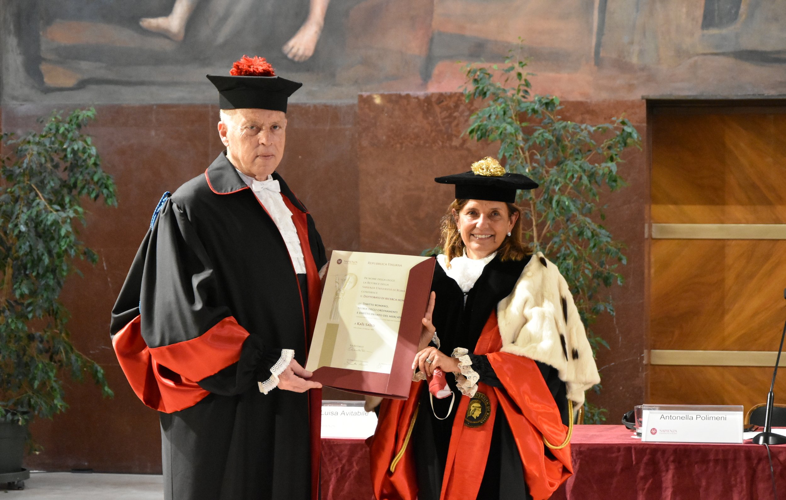 L'immagine mostra due persone vestite in abiti accademici formali, tipici per cerimonie di laurea o conferimenti di titoli. Il soggetto a sinistra indossa un mantello nero con dettagli rossi e un cappello, mentre a destra c'è una donna con un abito simile, ma con un mantello e un cappello che contengono diversi elementi decorativi. Entrambi stanno tenendo un documento o un certificato che sembra essere di rilevanza accademica o ufficiale. Sullo sfondo, ci sono piante e un muro decorato, indicando un ambiente formale e celebrativo.