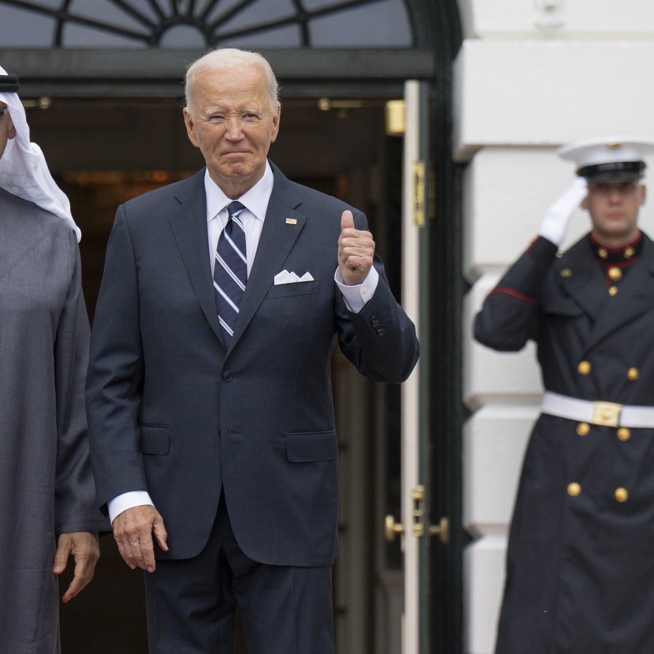 The image shows two men standing at the entrance of a building, likely the White House. One man is dressed in traditional attire with a white headscarf, while the other is in a formal suit. They are both smiling and posing for the camera, with one man waving and the other giving a thumbs-up. In the background, there are uniformed guards standing at attention. The scene conveys a sense of diplomacy and cordiality between the two leaders.