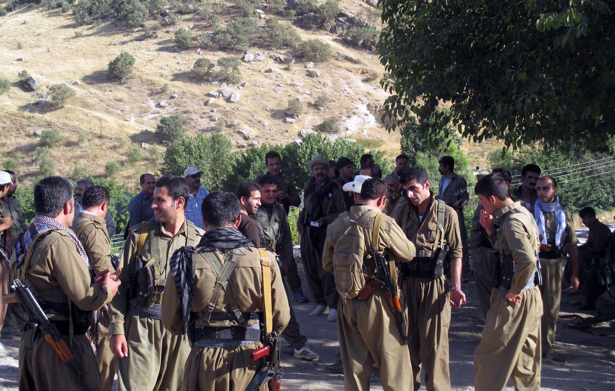 L'image montre un groupe de personnes rassemblées dans un environnement rural, avec des collines verdoyantes en arrière-plan. Les individus portent des vêtements traditionnels et certaines personnes sont armées. Ils semblent engager dans des discussions, tandis que d'autres se rassemblent autour d'eux. La scène reflète une atmosphère communautaire en milieu naturel.