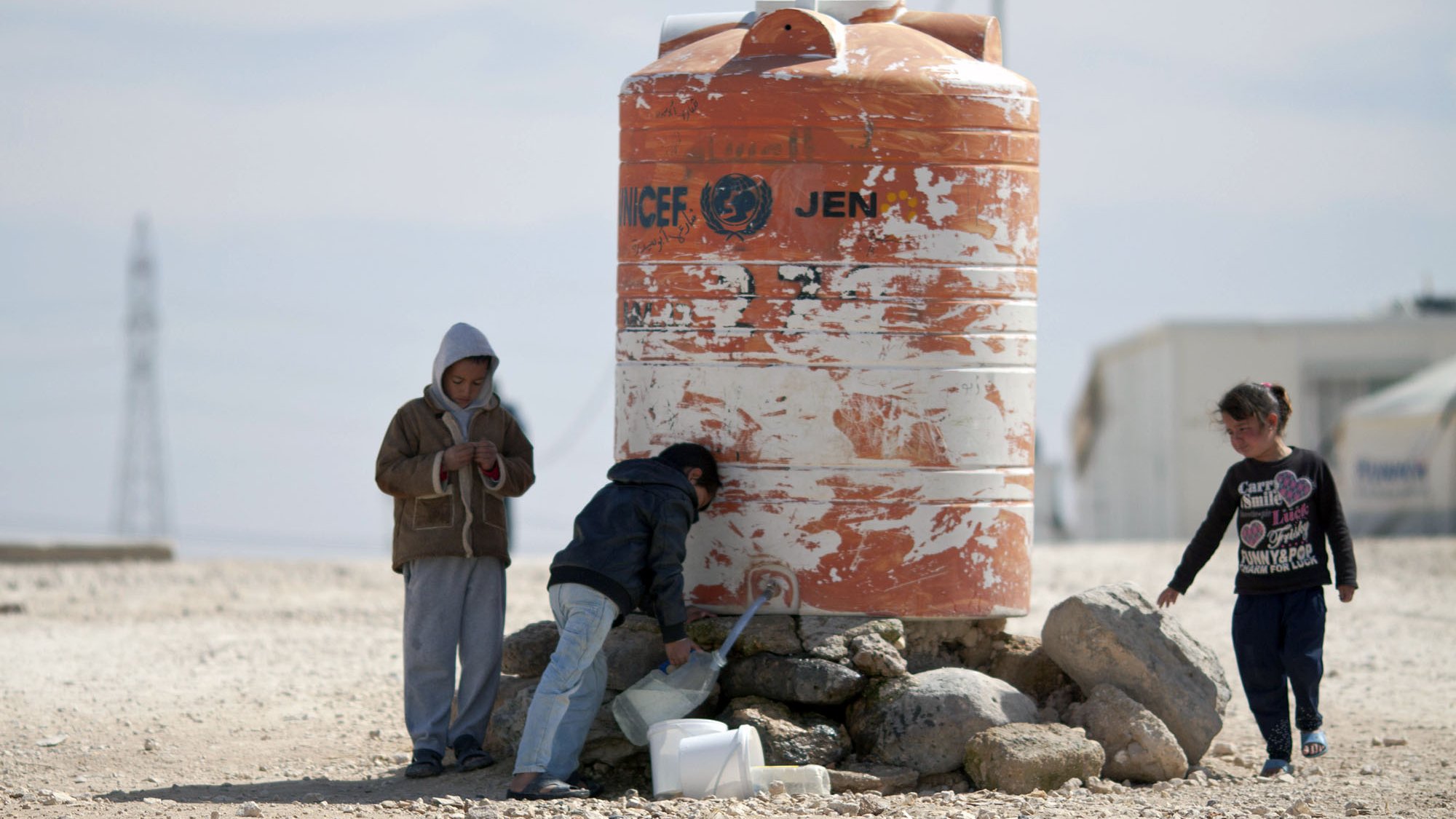 The image depicts children interacting near a large water tank. One child appears to be filling a container with water from a tap on the tank, while another child stands beside them. A third child is standing apart, looking at their phone or device. The water tank is orange and white, marked with a logo that indicates it is associated with a humanitarian organization. The surrounding area is dry and rocky, suggesting a challenging environment.
