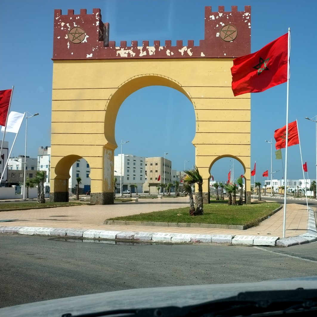 L'image montre un grand arc de triomphe peint en jaune avec des éléments rouges en haut, au centre d'une route. Drapeaux marocains flottent de chaque côté de l'arc, ajoutant une touche nationale à la scène. Des bâtiments modernes peuvent être aperçus au loin, tandis que la route est bordée de palmiers et d'une pelouse bien entretenue. Le ciel est clair et bleu, ce qui donne une ambiance ensoleillée et agréable à l'ensemble.