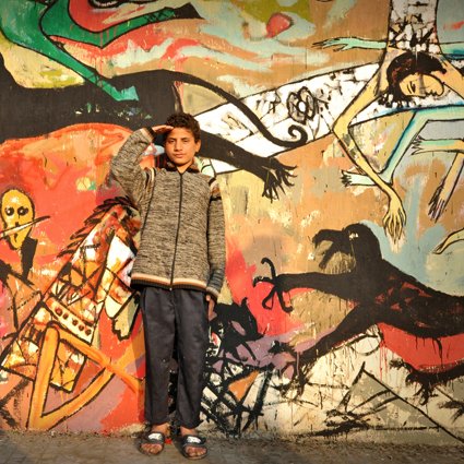 L'image montre un jeune garçon en train de faire un salut militaire devant un mur coloré, sur lequel sont peintes diverses figures stylisées et abstraites. Le mur présente des scènes dynamiques, avec des motifs de monstres et d'êtres humains, créant un contraste frappant avec le garçon qui porte un manteau gris et un pantalon sombre. Son expression et sa posture semblent témoigner de confiance ou de détermination. Les couleurs vives et les formes exagérées de la fresque attirent l'œil et ajoutent une dimension artistique à la scène.