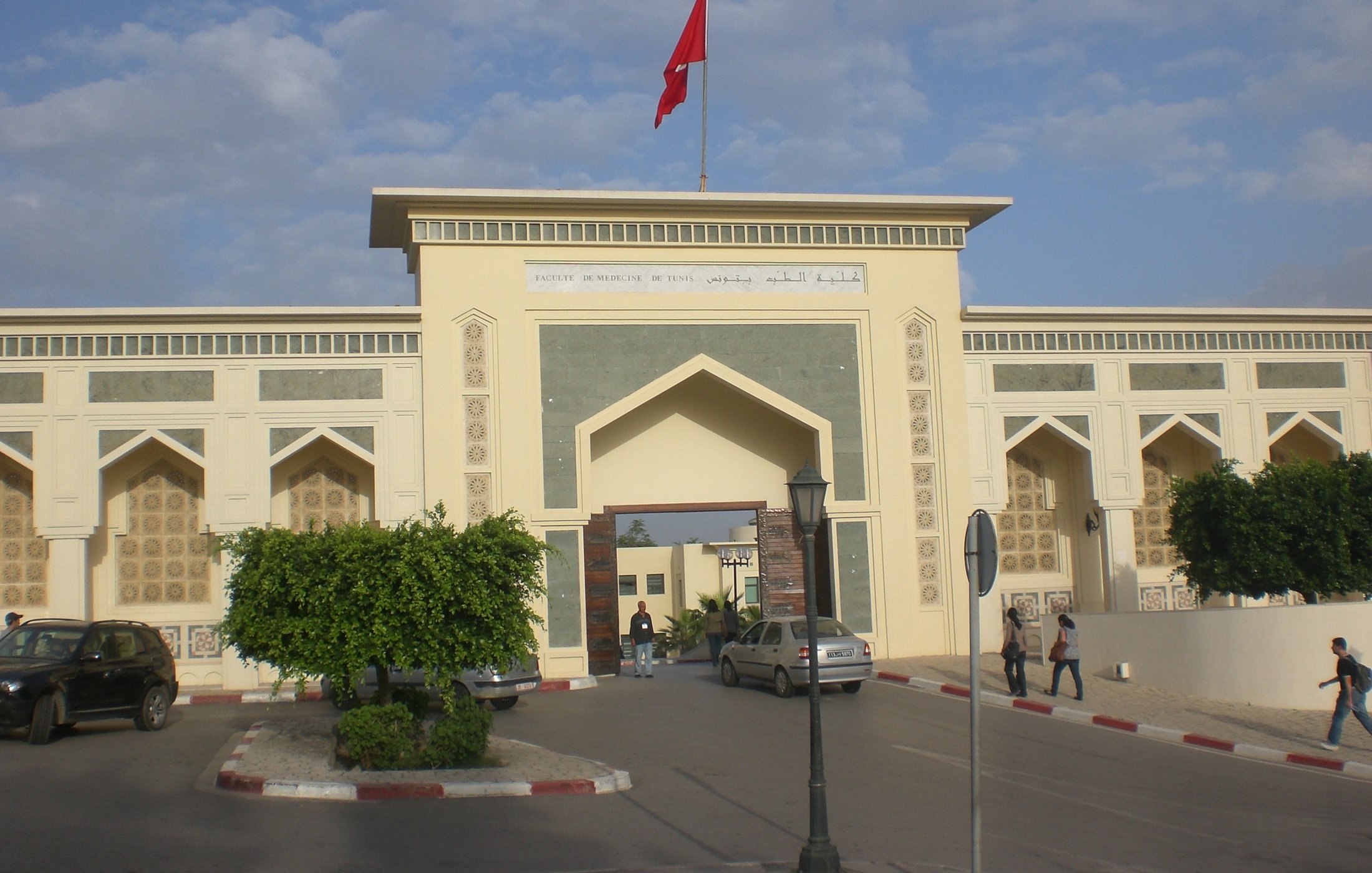 L'image montre l'entrée d'un bâtiment impressionnant, probablement une institution ou un musée, avec une architecture marocaine caractéristique. On peut voir une grande porte ornée de motifs traditionnels et des murs décorés avec des inscriptions en arabe et en français. Au-dessus de l'entrée, un drapeau rouge flotte. L'environnement est urbain, avec quelques arbres et des voitures stationnées. Le ciel est partiellement nuageux, apportant une atmosphère calme.