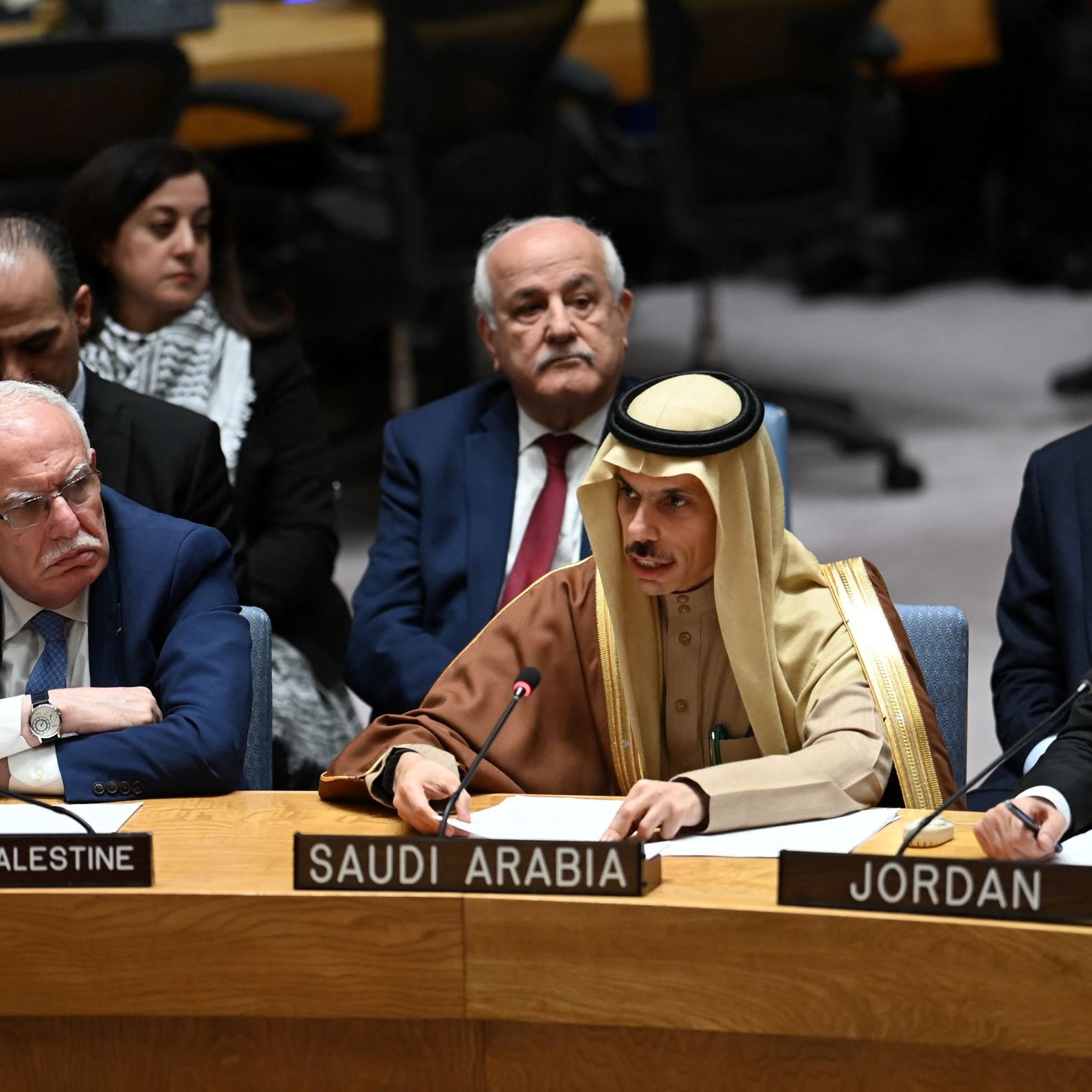 L'image montre une réunion de l'Assemblée générale ou du Conseil de sécurité de l'ONU. Plusieurs représentants de différents pays sont assis autour d'une table. À gauche, on voit un homme portant des lunettes avec un badge indiquant "État de Palestine". Au centre, un homme en tenue traditionnelle saoudienne prend la parole, tandis qu'un autre à droite, en costume noir, représente la Jordanie. En arrière-plan, d'autres participants écoutent attentivement. L'atmosphère semble sérieuse, suggérant une discussion importante.