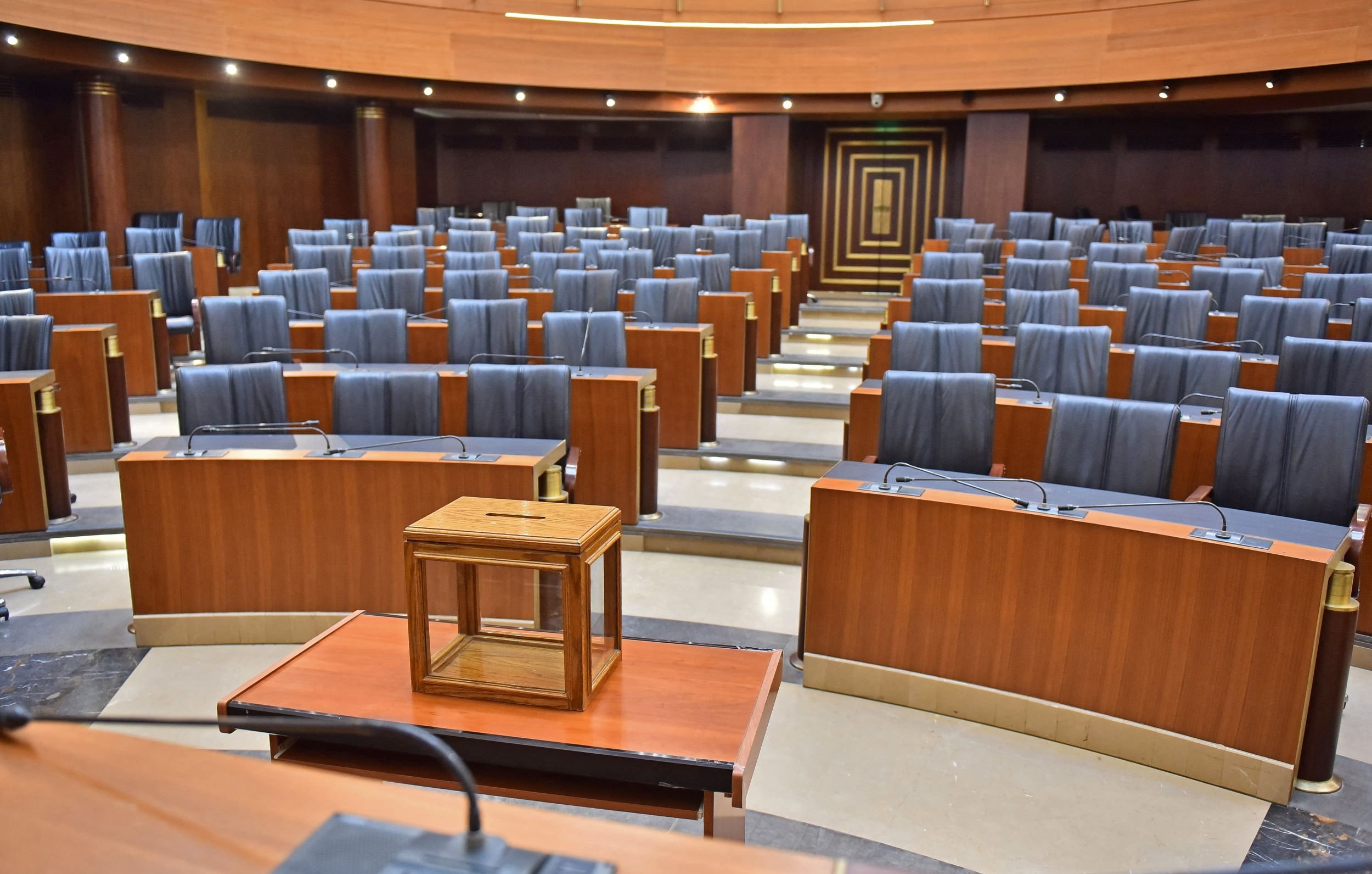 L'immagine mostra un'aula legislativa vuota, caratterizzata da una disposizione ordinata di banchi in legno con sedute nere. Al centro della scena si trova una piccola struttura in legno, probabilmente un tavolo o una cassa. Le pareti dell'aula sono di un colore scuro, mentre il pavimento è di tonalità più chiara, creando un contrasto. L'ambiente sembra formale e istituzionale, suggerendo un luogo di discussione e decisione politica.
