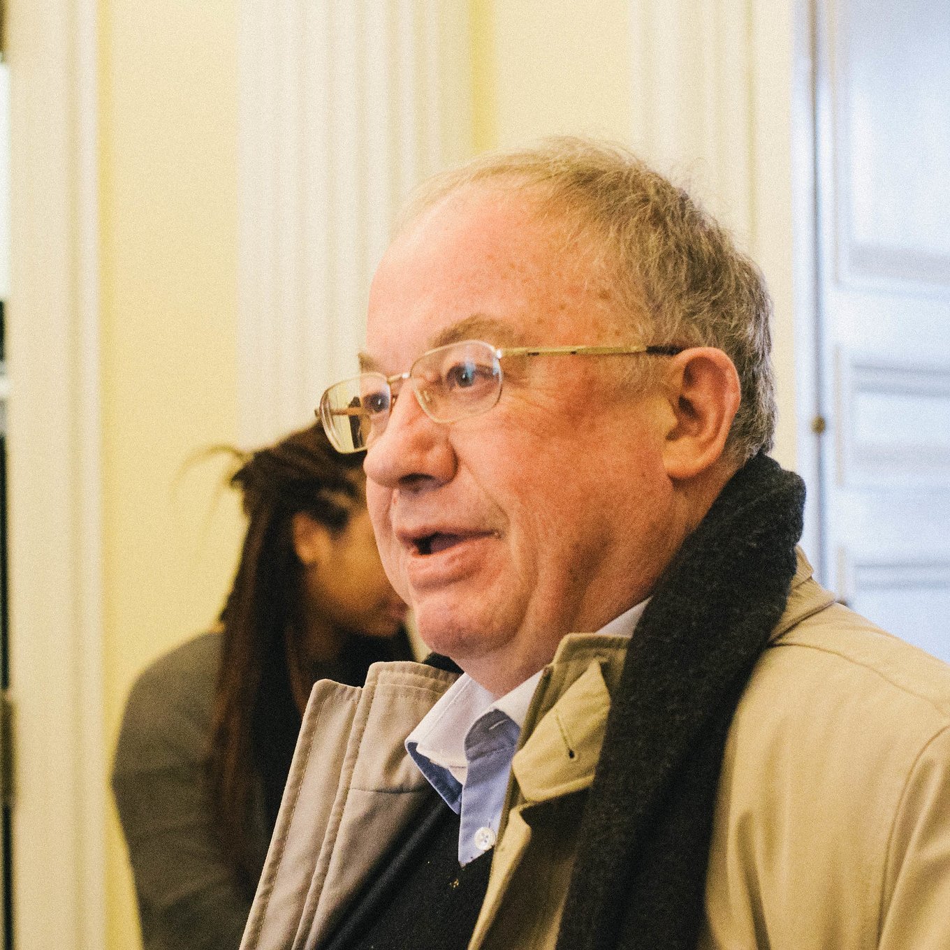 Homme âgé avec des lunettes, portant un manteau, dans un intérieur lumineux.