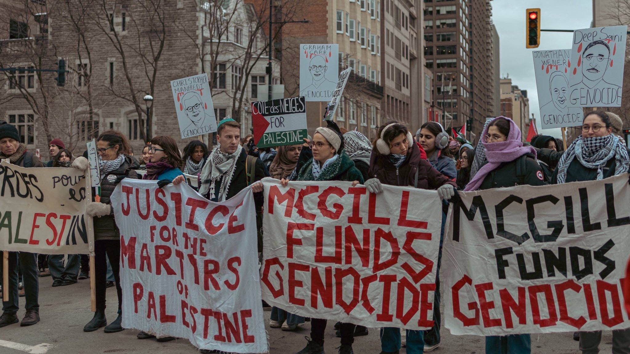 L'image montre une manifestation dans une ville urbaine. Un groupe de personnes est rassemblé, tenant des pancartes avec des messages en faveur de la Palestine et s'opposant au financement de ce qu'ils appellent un génocide. Certains manifestants portent des écharpes qui semblent indiquer leur solidarité avec la cause palestinienne. L'atmosphère est engagée et déterminée, avec des passants et des bâtiments en arrière-plan.