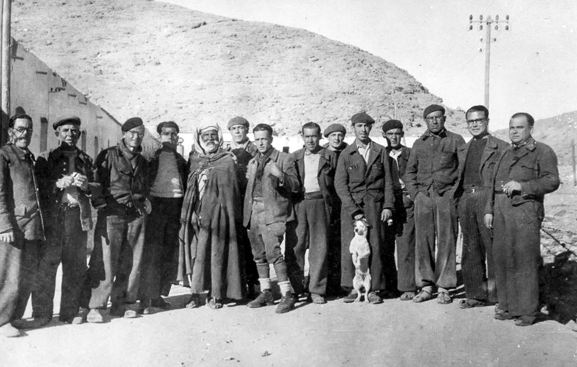 L'image en noir et blanc montre un groupe d'hommes se tenant debout ensemble, probablement dans un environnement rural ou montagneux. Ils portent des vêtements qui semblent être des tenues de travail ou militaires. Au centre, un homme en costume traditionnel se distingue des autres. Un chien se trouve également parmi le groupe. Les hommes affichent diverses expressions, allant des sourires aux regards sérieux. L'arrière-plan présente des collines, et quelques bâtiments peuvent être aperçus. L'ensemble évoque une période historique, probablement au début ou au milieu du 20e siècle.