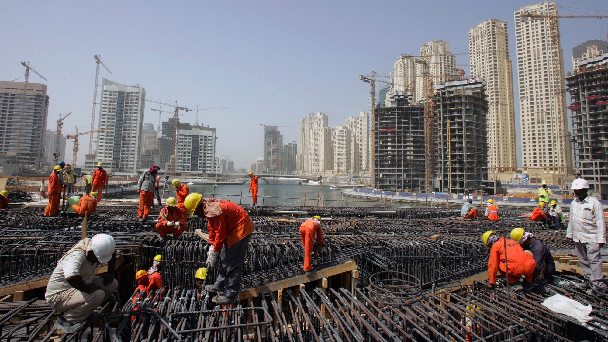 الصورة تظهر موقع بناء في منطقة حضرية حديثة. يتواجد فيها العديد من العمال يرتدون ملابس العمل البرتقالية والخوذ البيضاء. تشير الصورة إلى أن العمال مشغولون بعمليات البناء، حيث يمكن رؤية الهياكل الحديدية والرافعات في الخلفية. المباني الشاهقة الحديثة المحيطة بالموقع تدل على تطور المدينة، مع وجود بعض العمارات قيد الإنشاء. السماء تبدو صافية، ولكن هناك غبار خفيف يوحي بنشاط البناء.