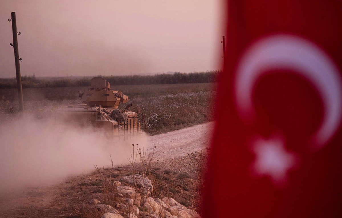L'image montre un tank, probablement militaire, se déplaçant sur une route poussiéreuse. Au premier plan, on aperçoit un drapeau turc, qui ajoute un contexte national à la scène. L'arrière-plan présente un paysage rural, avec des champs, et l'on peut voir de la poussière se lever autour du tank, suggérant une atmosphère de mouvement ou d'activité militaire. L'éclairage est doux, créant une ambiance de crépuscule.