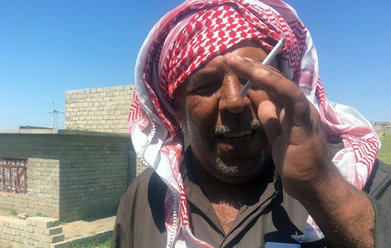 L'image montre un homme portant une tenue traditionnelle avec un keffieh sur la tête. Il semble être à l'extérieur, avec des bâtiments en arrière-plan. L'homme fume une cigarette et sourit légèrement en regardant vers la caméra. Le paysage semble rural, avec de l'herbe et des structures en pierre.