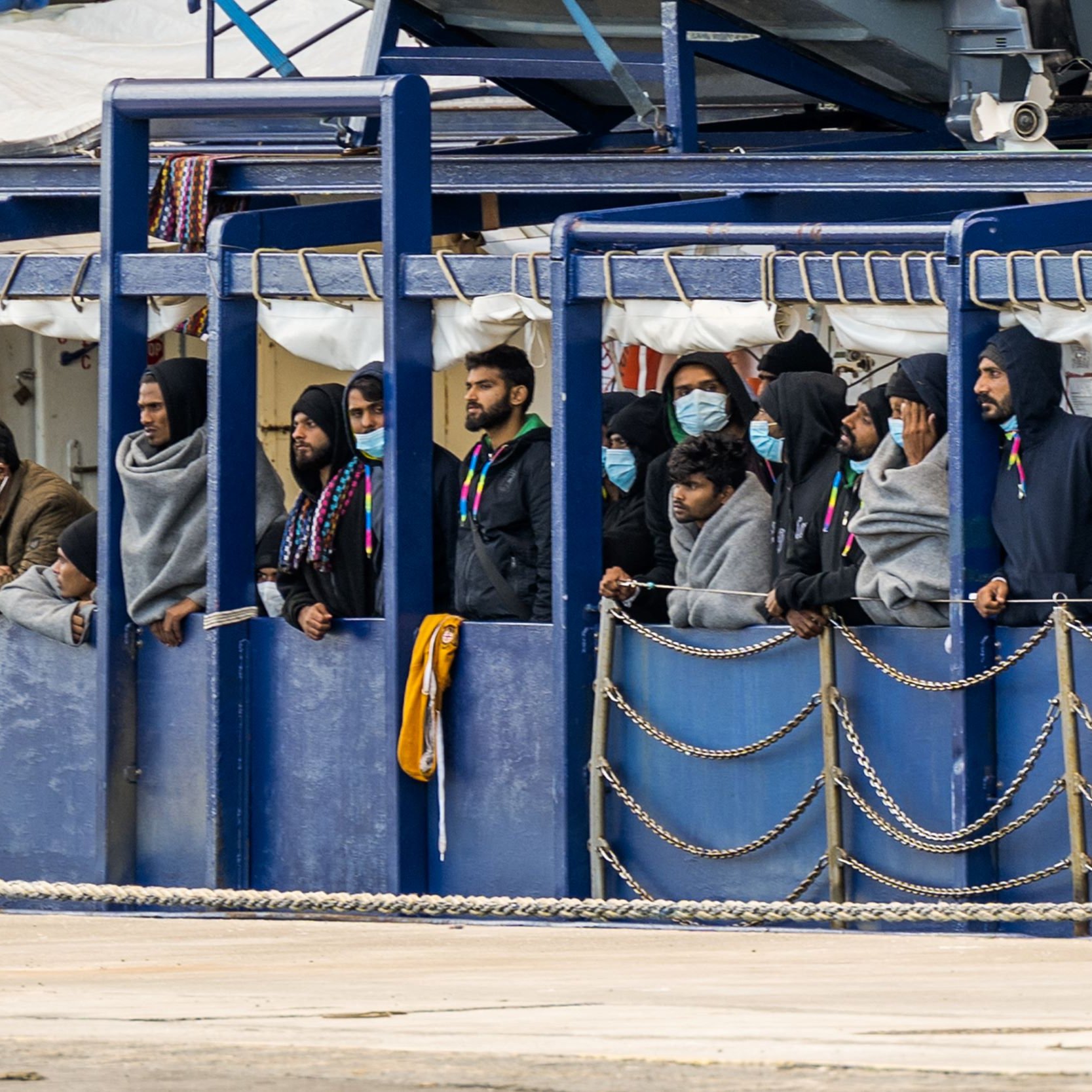 در تصویر، گروهی از افراد در یک محیط بسته مشغول تماشا هستند. آن‌ها ماسک‌های بهداشتی به چهره دارند و برخی از آن‌ها لباس‌های گرم و کلاه بر سر دارند. به نظر می‌رسد که آن‌ها در کنار یک کشتی یا قایق ایستاده‌اند و به چیزی در دوردست نگاه می‌کنند. محیط اطراف شامل سازه‌هایی از فلز و موانع زنجیری است. احساس صمیمیت و توجه در چهره‌هایشان دیده می‌شود.