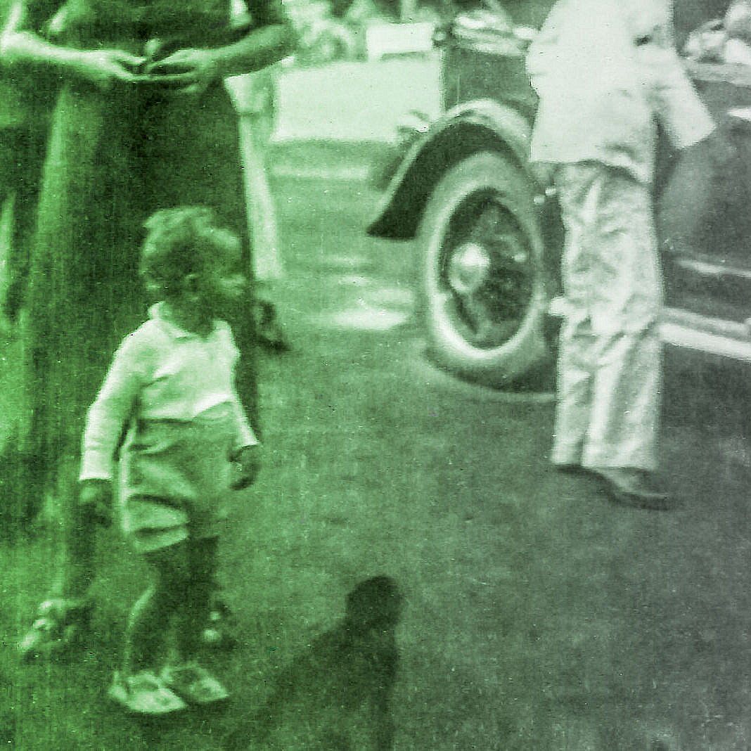 L'image montre un groupe de personnes en extérieur, probablement dans un cadre informel ou festif. On peut voir un enfant en bas à gauche, habillé en short, qui semble jouer. Des adultes, qui peuvent être des membres de la famille ou des amis, se tiennent près d'une voiture vintage, ce qui ajoute une touche rétro à la scène. L'atmosphère semble conviviale et joyeuse. Les vêtements et le style des personnages évoquent une époque passée.