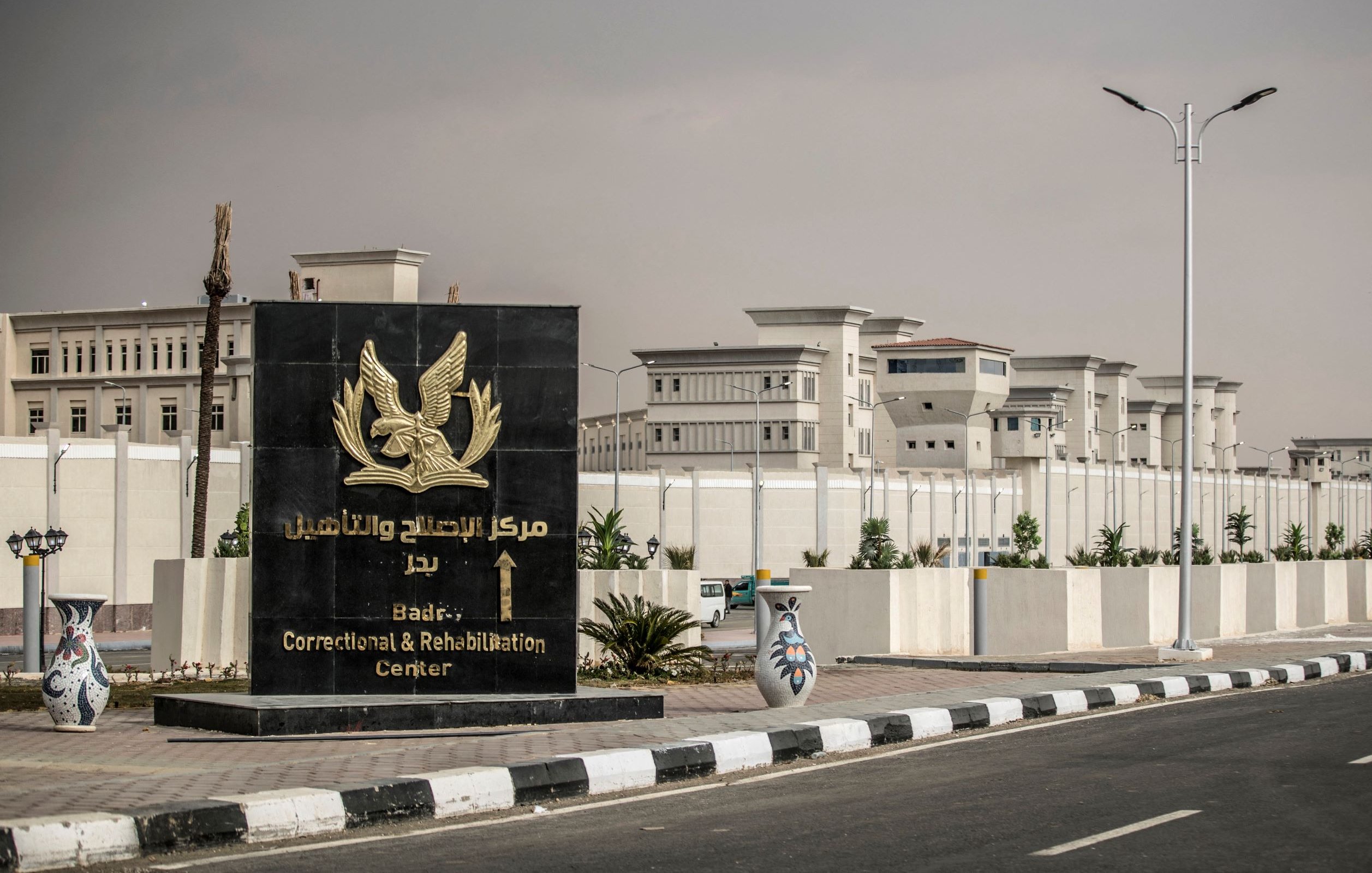 L'image montre un grand panneau d'entrée d'un centre de correction et de réhabilitation, situé à Badri, en arabe et en anglais. En arrière-plan, on peut voir des bâtiments modernes, probablement liés à ce centre. L'environnement semble urbain, avec des routes pavées et des éléments paysagers tels que des plantes. Le ciel semble légèrement nuageux, donnant une ambiance un peu sombre à la scène.