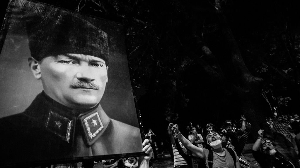 L'image montre un rassemblement nocturne où des gens tiennent un grand portrait d'une figure historique. Les participants semblent exprimer leur admiration ou leur respect, levant le poing en signe de solidarité ou de protestation. L'atmosphère est festive mais aussi solennelle, avec des ombres et une luminosité faible qui accentue l'intensité du moment.