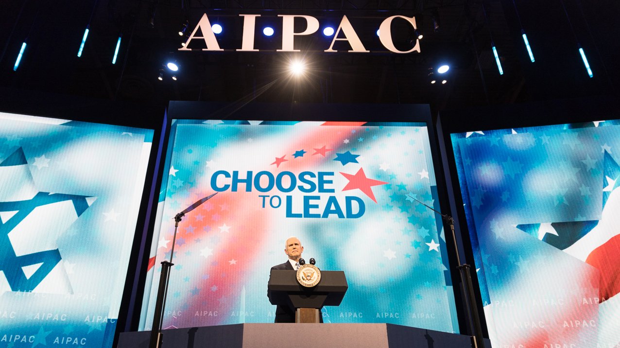 تظهر الصورة منصة خطابة كبيرة تحمل شعار "AIPAC" في الخلفية. يتواجد متحدث خلف ميكروفون على المنصة، مع وجود شاشات عرض كبيرة على الجانبين تظهر عبارة "اختَر القيادة" مع نجوم وشعارات ذات طابع إسرائيلي. الإضاءة تركز على المتحدث، مما يبرز أهمية الحدث الذي يُعقد.