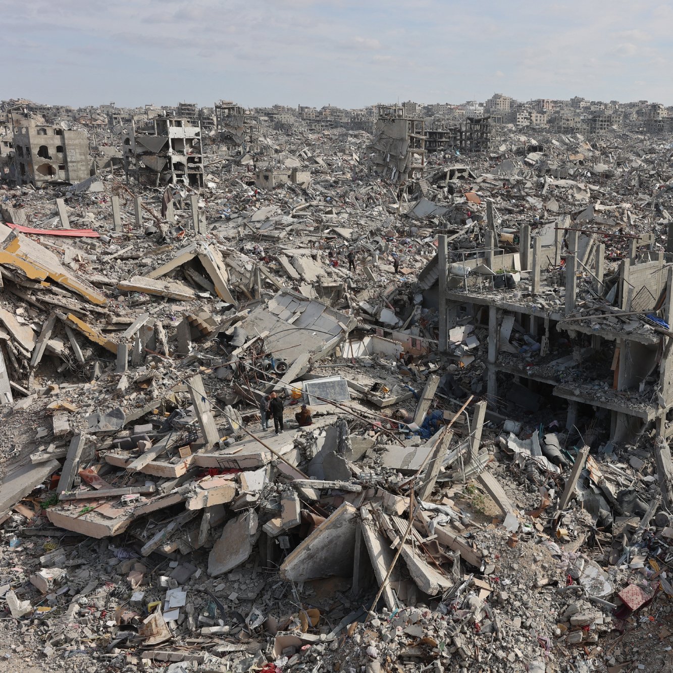 تصویر یک منطقه خرابه است که در آن ساختمان‌ها و سازه‌های ویران‌شده به چشم می‌خورند. زمین پوشیده از بتن و مصالح ساختمانی است و فضایی خالی و تکان‌دهنده دارد. صدای باد و سکوت سنگین در این محیط حاکم است.