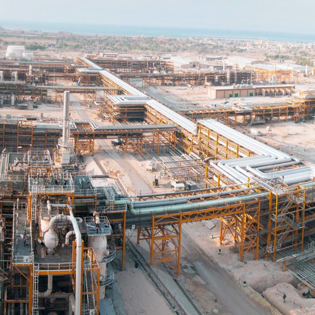 الصورة تُظهر مشهدًا واسعًا لمجمع صناعي، حيث تظهر مرافق ومعدات ضخمة. يمكن رؤية أنابيب كبيرة تمتد عبر الموقع، بالإضافة إلى أفران ومنشآت أخرى تشير إلى نشاط صناعي مكثف. الخلفية تتضمن المساحات المفتوحة والمباني التي تدل على طبيعة العمل في هذا المكان. السماء تبدو صافية، مما يضفي طابعًا مشمسًا على الصورة.