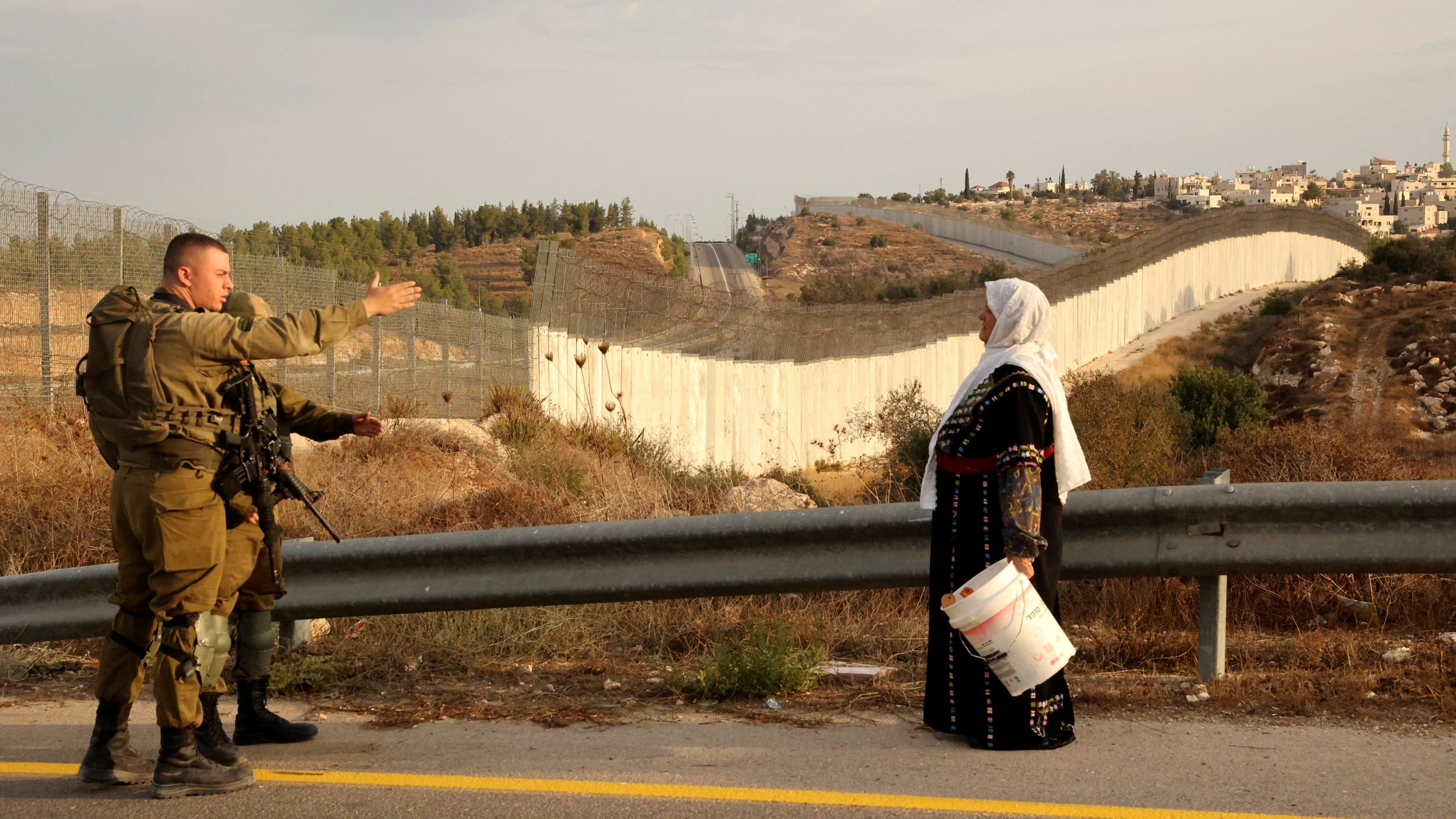 L'image montre un soldat en uniforme qui semble communiquer avec une femme vêtue de manière traditionnelle, tenant un seau. En arrière-plan, on aperçoit un mur ou une barrière qui sépare les deux côtés, avec des collines et des bâtiments au loin. L'environnement semble rural, avec de la végétation et une route. Cette scène évoque un contexte de conflit ou de tension dans une région marquée par des divisions.