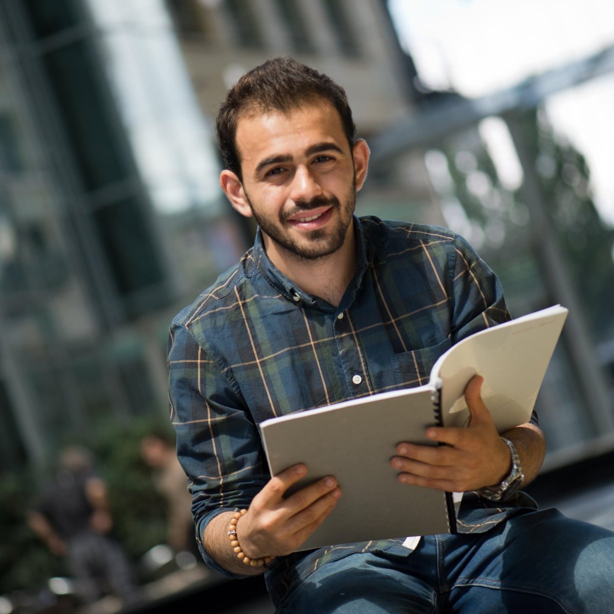 L'image montre un jeune homme assis sur un banc, souriant, tout en tenant un carnet ou un cahier dans ses mains. Il porte une chemise à carreaux et a l'air détendu, avec un environnement urbain en arrière-plan et des bâtiments modernes. La lumière naturelle éclaire la scène, créant une atmosphère agréable.