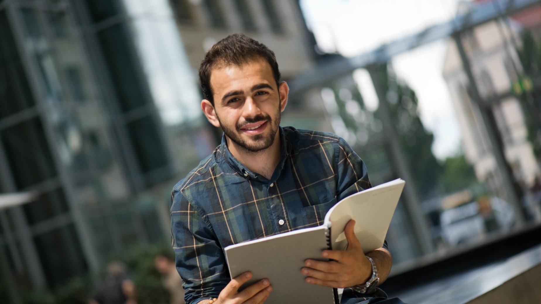 The image features a young man sitting outdoors, holding an open notebook or binder. He has a friendly smile and is wearing a plaid shirt. The background includes modern architecture and greenery, suggesting an urban setting. The atmosphere appears bright and inviting, indicating a pleasant day.