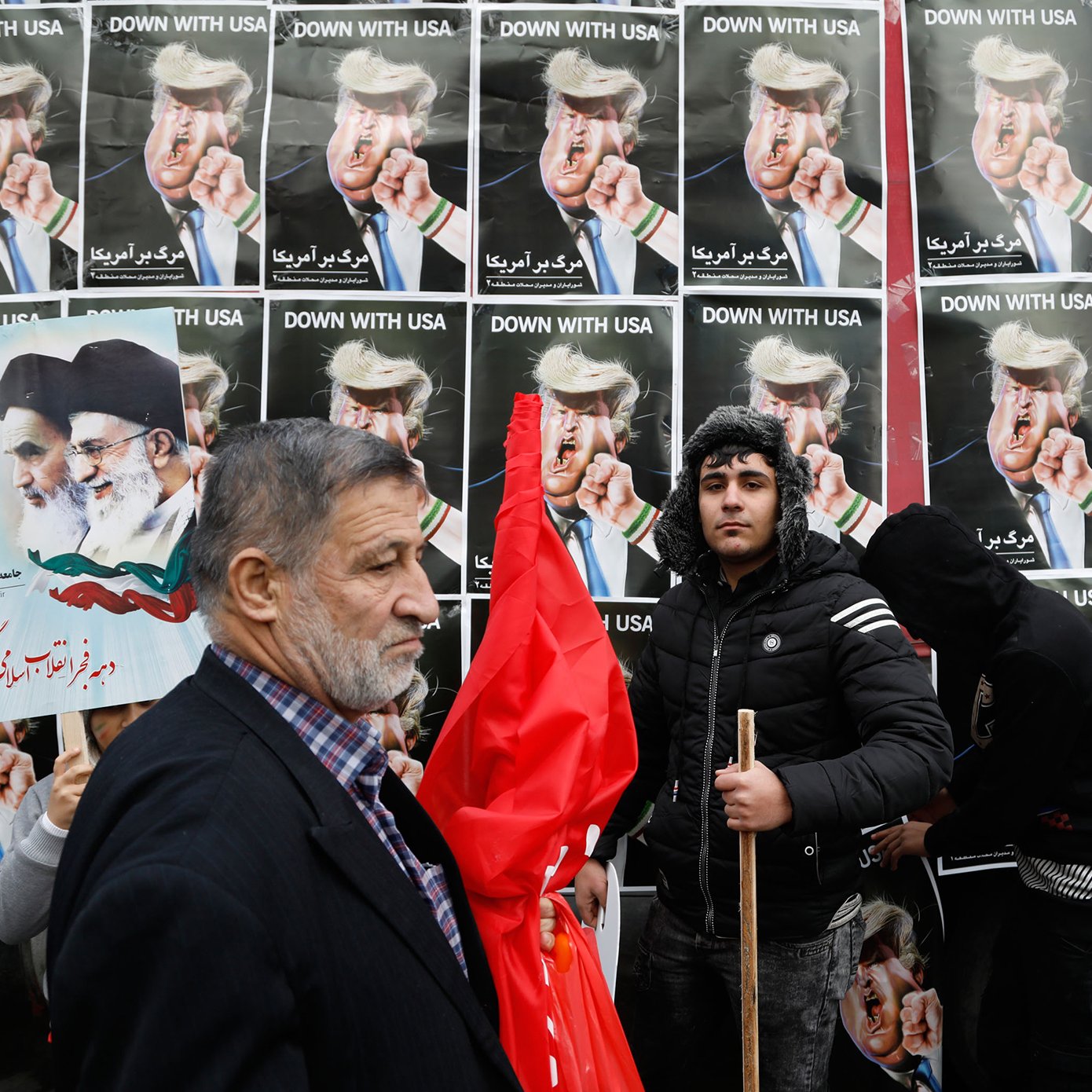 در این تصویر، گروهی از مردم در حال تظاهرات به نمایش گذاشته‌اند. در پس‌زمینه، پوسترهایی با تصاویر رهبران سیاسی و متنی به زبان فارسی دیده می‌شود که به موضوعی خاص اشاره دارد. یکی از افراد به سمت دوربین نگاه می‌کند و فرد دیگری با یک چوب در دست و لباسی سیاه در کنار او ایستاده است. برخی از افراد دیگر نیز در این صحنه حضور دارند که نشان‌دهنده جو تظاهرات است. پرچم‌ها و پلاکاردها نیز در میان جمعیت دیده می‌شود که نشان‌دهنده احساسات و پیام‌های سیاسی آن‌ها است.