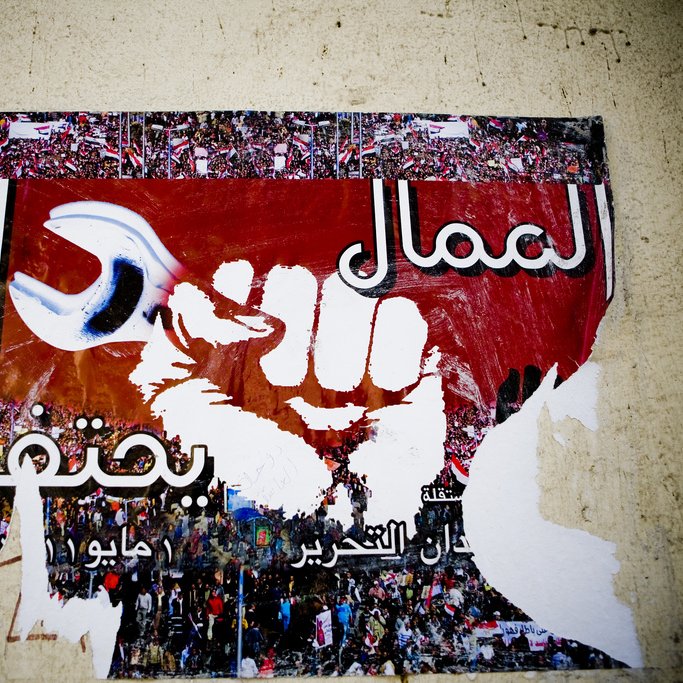 L'image montre une affiche partiellement déchirée qui présente un poing levé, symbole de révolte ou de solidarité. En arrière-plan, on peut apercevoir une grande foule, probablement lors d'une manifestation. Le fond est rouge, et il y a des inscriptions en arabe et en d'autres langues qui semblent exprimer des messages politiques ou sociaux. Le tout évoque un fort sentiment de lutte collective et de volonté de changement.