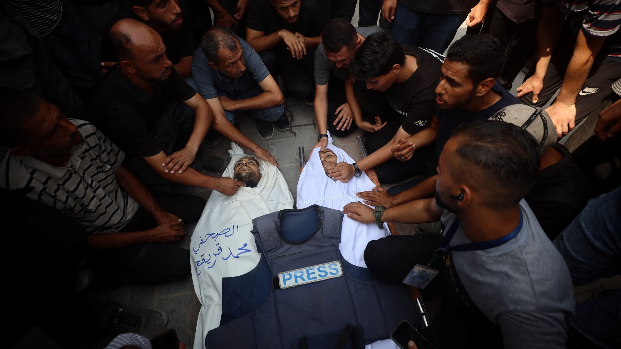 A crowd mourns over a deceased person covered in a white cloth, wearing a press vest.
