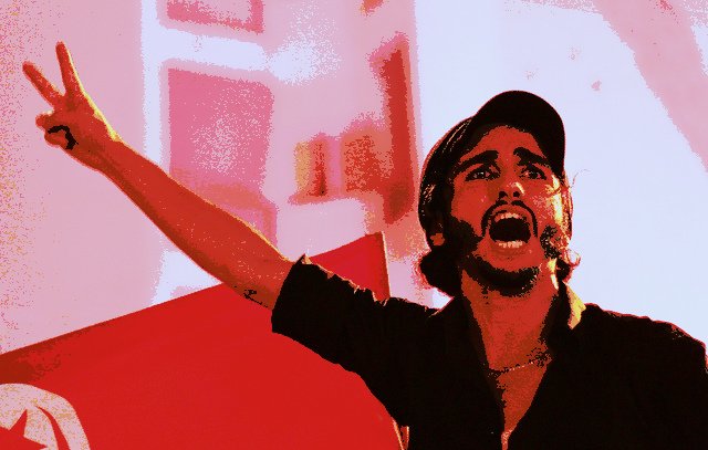 L'image montre un homme debout, apparemment engagé dans une expression de passion ou de colère. Il lève son bras en faisant un signe de victoire. Son visage est très expressif, avec une bouche ouverte comme s'il était en train de crier ou de s'exprimer avec force. En arrière-plan, on peut discerner un drapeau, qui pourrait suggérer un contexte politique ou social. Les couleurs de l'image sont assez vives, ce qui accentue l'intensité de la scène.