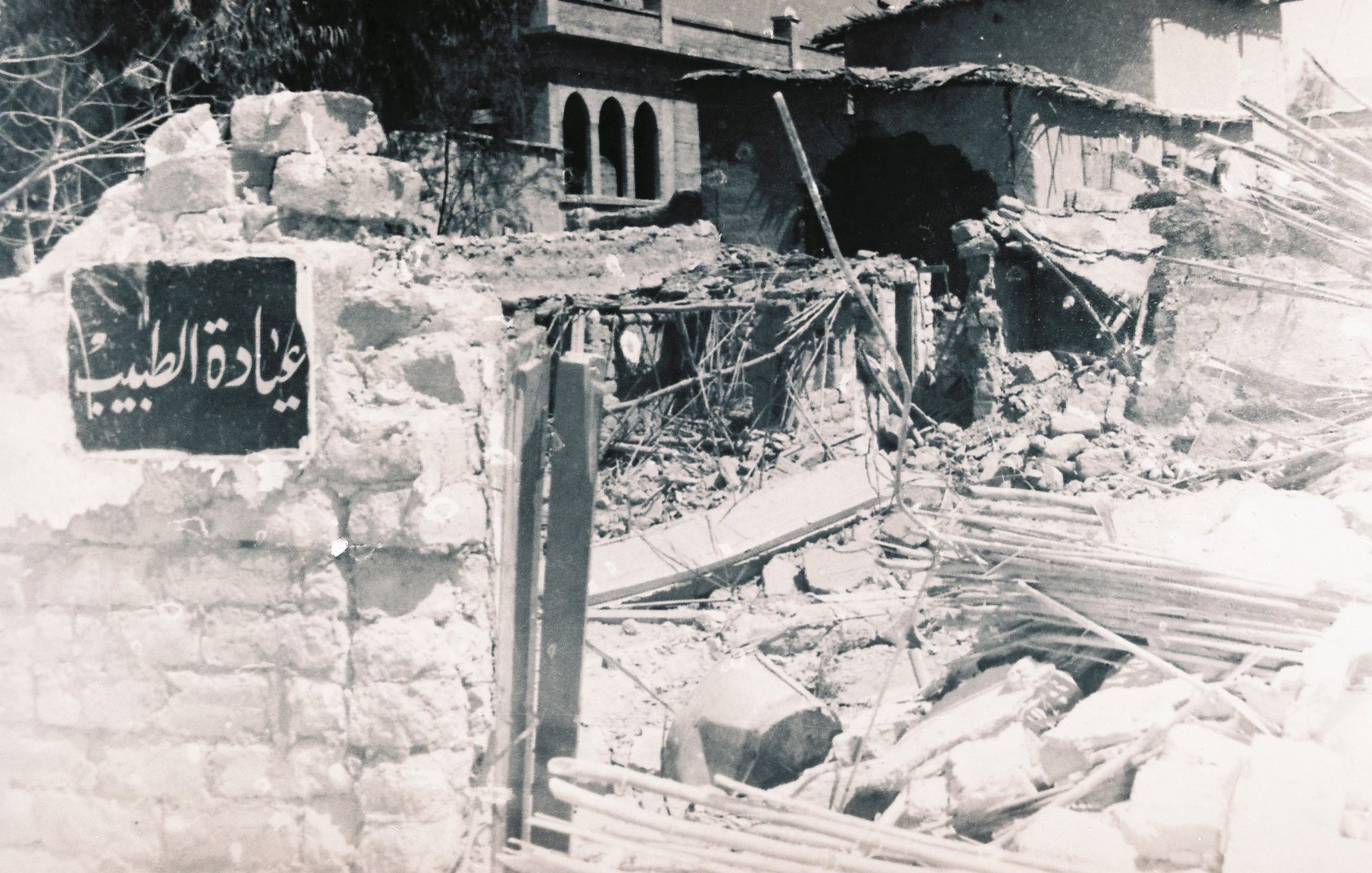 الصورة تُظهر مشهداً لمدمرات بسبب أحداث سابقة، حيث يظهر حطام المباني والخرائب. في مقدمة الصورة، توجد لافتة تحمل اسم مكان، ولكنها مهملة وسط الأنقاض. الخلفية تحتوي على نوافذ وأجزاء من مباني قديمة، مما يعكس تأثير الزمن أو كارثة معينة. الأجواء توحي بالحزن والألم الناتج عن الدمار.