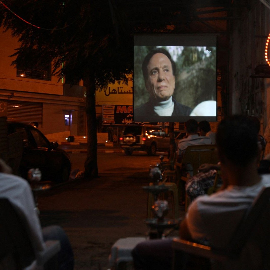 L'image montre une scène nocturne où un groupe de personnes est assis à l'extérieur, observant un film projeté sur un mur. On voit des chaises disposées face à l'écran, et quelques fumées de narguilé flottent dans l'air. L'environnement est animé par des lumières de néon, et on aperçoit des voitures garées et des bâtiments en arrière-plan, créant une ambiance conviviale et détendue.