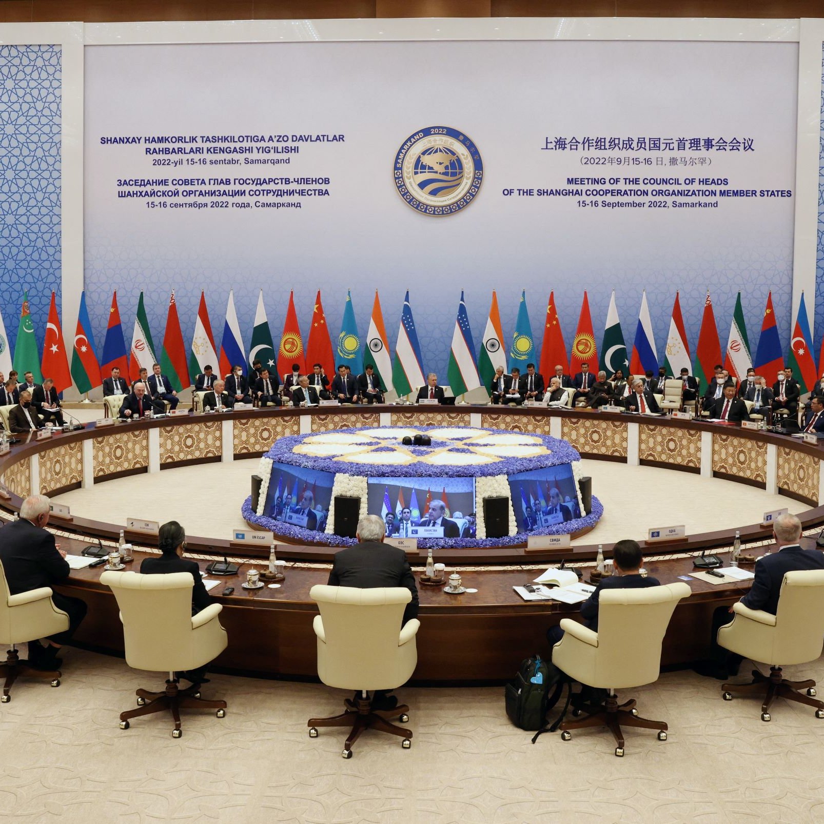 تظهر الصورة اجتماعاً رسمياً حيث يجلس عدد من القادة والممثلين حول طاولة دائرية كبيرة. يتميز المكان بزينة أنيقة، والجدران مزينة بأعلام دول مختلفة. يبدو أن هذا الاجتماع يهدف إلى مناقشة قضايا مهمة، حيث يظهر المشاركون في شكل جاد ومركز. تمثل الطاولة الكبيرة مركز الاجتماع، بينما تتواجد لافتات تشير إلى الحدث والمكان الذي يُعقد فيه الاجتماع.