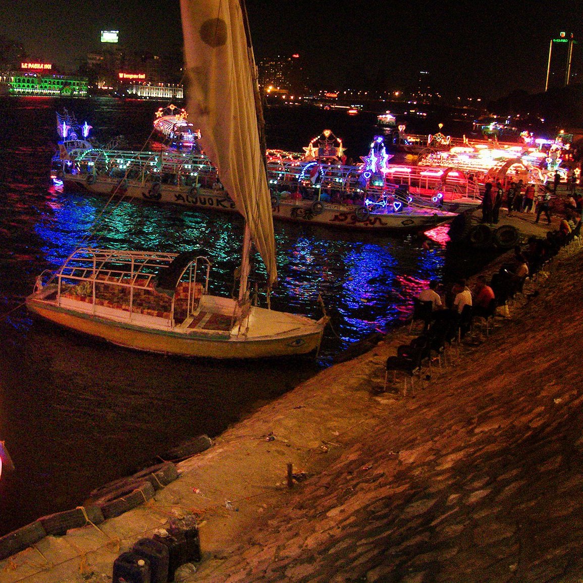 L'immagine mostra una scena notturna su un fiume, con diverse barche decorate con luci colorate. Le imbarcazioni si trovano in una zona animata, e il riflesso delle luci sull'acqua crea un'atmosfera vivace e festosa. Sullo sfondo, si possono vedere edifici illuminati, che contribuiscono ulteriormente all'ambiente festivo della scena. Alcune persone sono sedute lungo la riva, godendosi la vista e l'atmosfera.