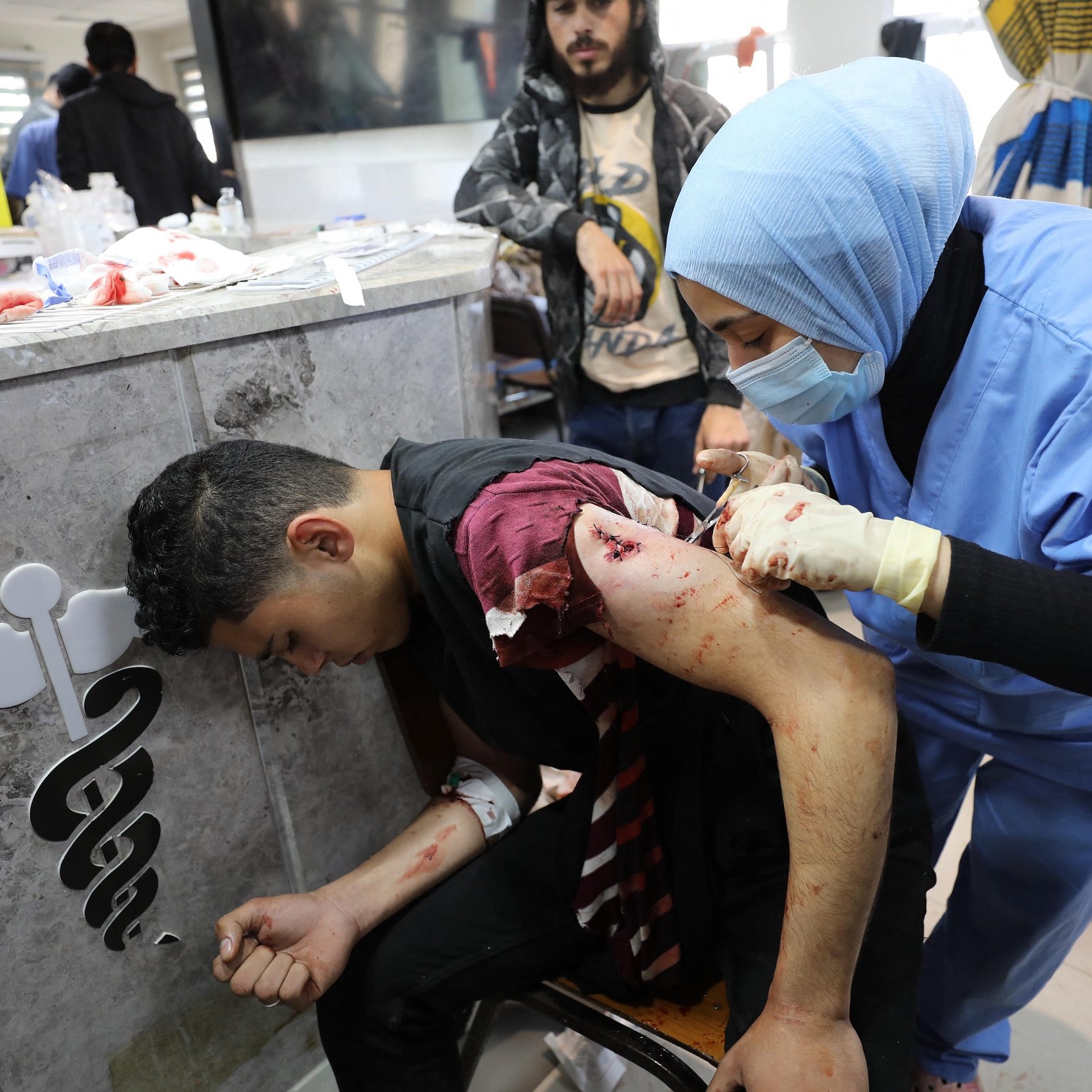 L'image montre une scène d'urgence médicale. Un jeune homme, assis sur une chaise, a des blessures visibles sur son bras et son épaule. Il semble souffrir et se penche en avant. À côté de lui, une professionnelle de la santé, vêtue d'une blouse bleue et portant un masque, traite ses blessures avec attention. L'environnement semble être un espace médical, avec des équipements et des fournitures autour. Des personnes attire également l'attention en arrière-plan, suggérant une situation de crise.