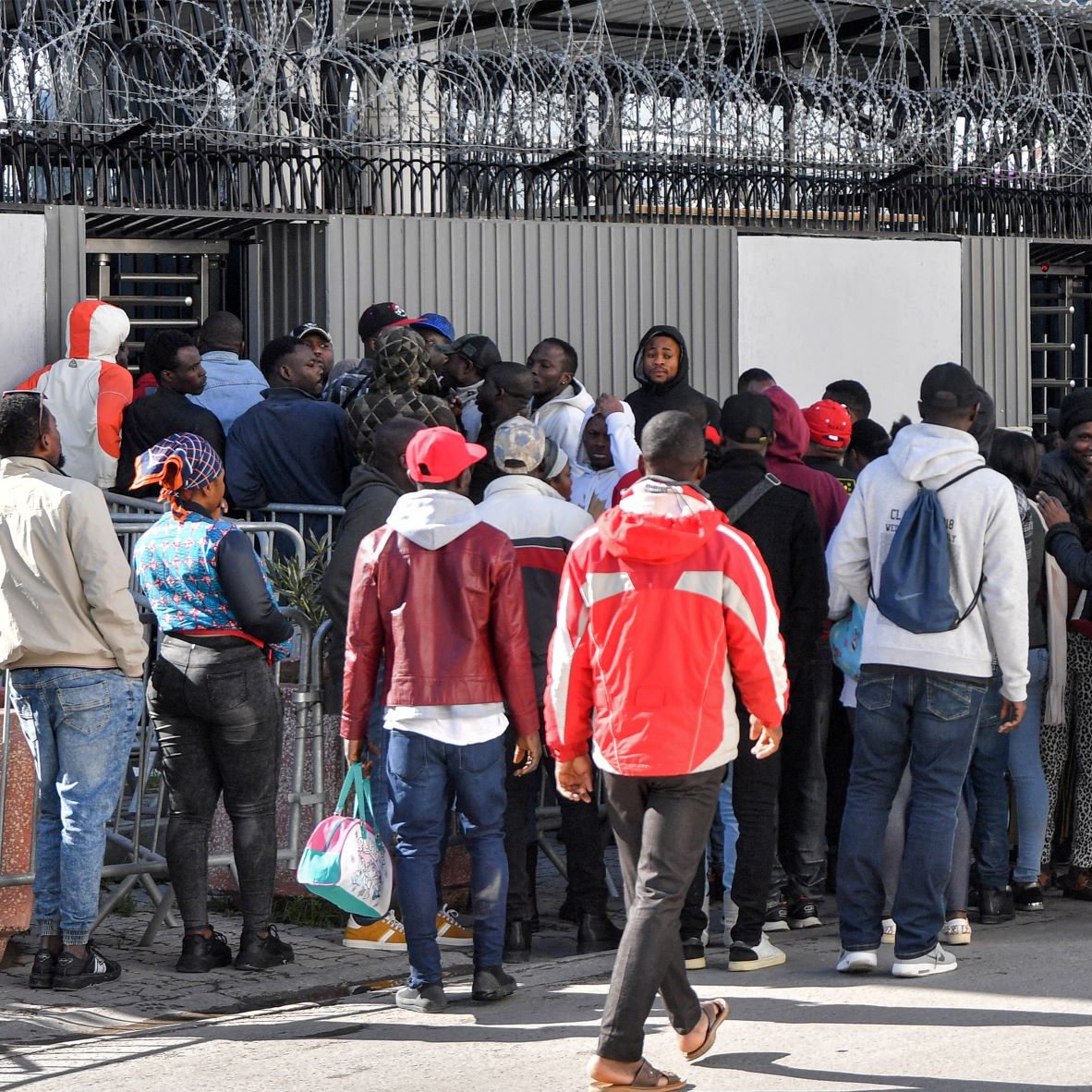 L'image montre un groupe de personnes rassemblées devant une clôture avec des barbelés. Elles semblent attendre ou essayer d'entrer dans un bâtiment. Certaines personnes portent des vêtements colorés, comme des vestes et des chapeaux rouges. Il y a une certaine densité dans la foule, suggérant une situation de rassemblement ou de file d'attente. Le mur en arrière-plan comporte des affichages et il y a une ambiance de tension ou d'importance autour de l'endroit où se trouvent les personnes.