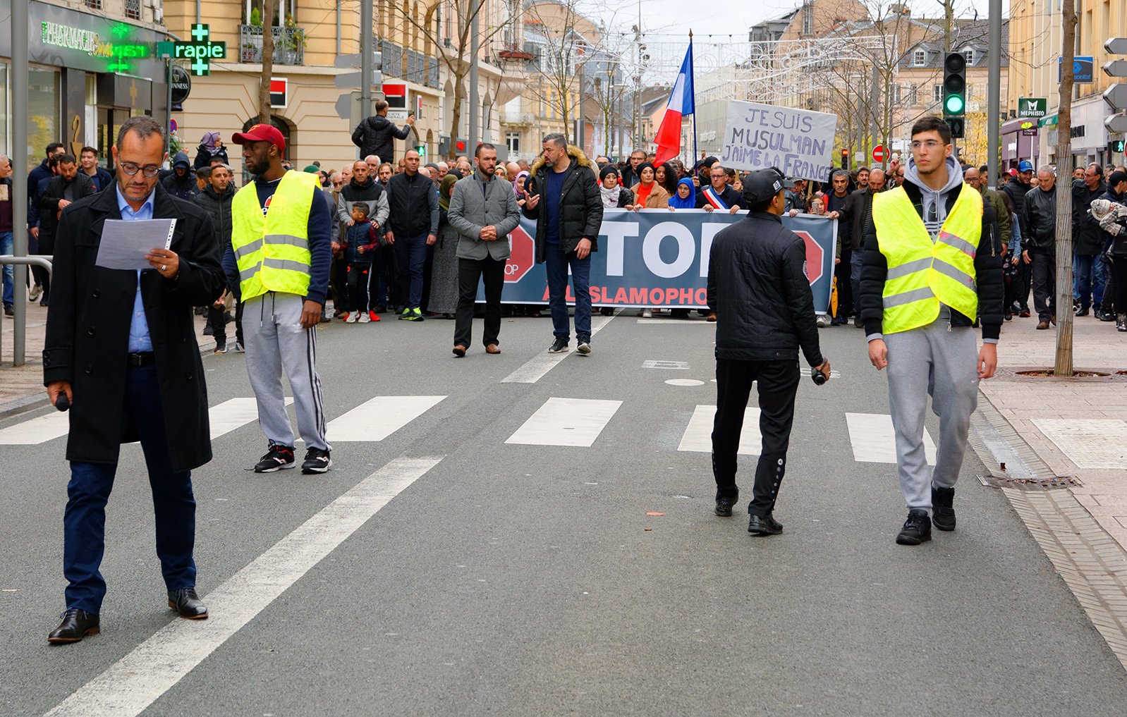L'image montre une manifestation dans une rue où un groupe de personnes tient une grande banderole avec des slogans. Au premier plan, deux agents de sécurité ou policiers portent des gilets réfléchissants et semblent surveiller la situation. Des manifestants sont visibles à l'arrière, et il y a une ambiance de mobilisation avec des drapeaux. L'environnement urbain se présente avec des bâtiments voisins et une circulation routière réduite à ce moment.