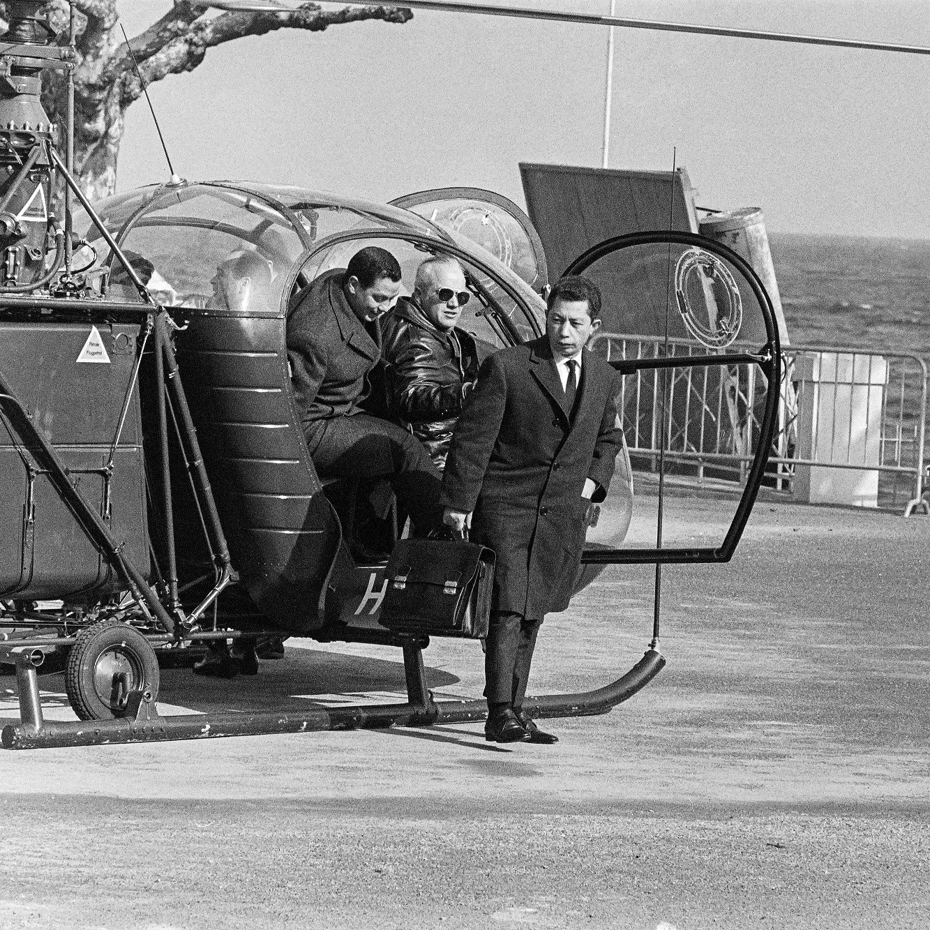 L'image montre une scène en noir et blanc avec un hélicoptère garé près de la mer. Deux personnes descendent de l'hélicoptère, tandis qu'une troisième personne les attend. On peut les voir porter des vêtements formels, ce qui suggère une certaine occasion. L'environnement est dégagé, avec une barrière visible en arrière-plan et des arbres dénudés. La mer se trouve en arrière-plan, ajoutant une touche de paysage côtier à la scène.