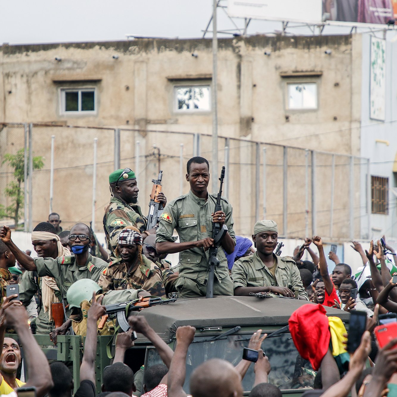 L'image montre un groupe de soldats, armés, sur un véhicule militaire, entourés par une foule enthousiaste. Les soldats semblent célébrer ou soutenir une cause, tandis que les membres de la foule lèvent les bras et expriment leur joie ou leur enthousiasme. En arrière-plan, on peut apercevoir des bâtiments et une atmosphère de mobilisation populaire.