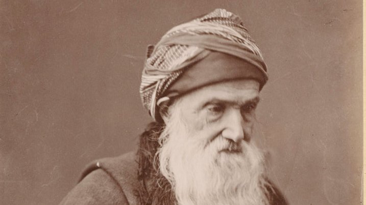 L'image montre un homme âgé avec une longue barbe blanche. Il porte un turban ou une coiffe enroulée autour de la tête, ainsi qu'une cape sombre. Son regard est concentré et il tient une canne. L'arrière-plan est neutre, mettant en valeur son visage et son expression, ce qui donne une impression de sagesse et de dignité.