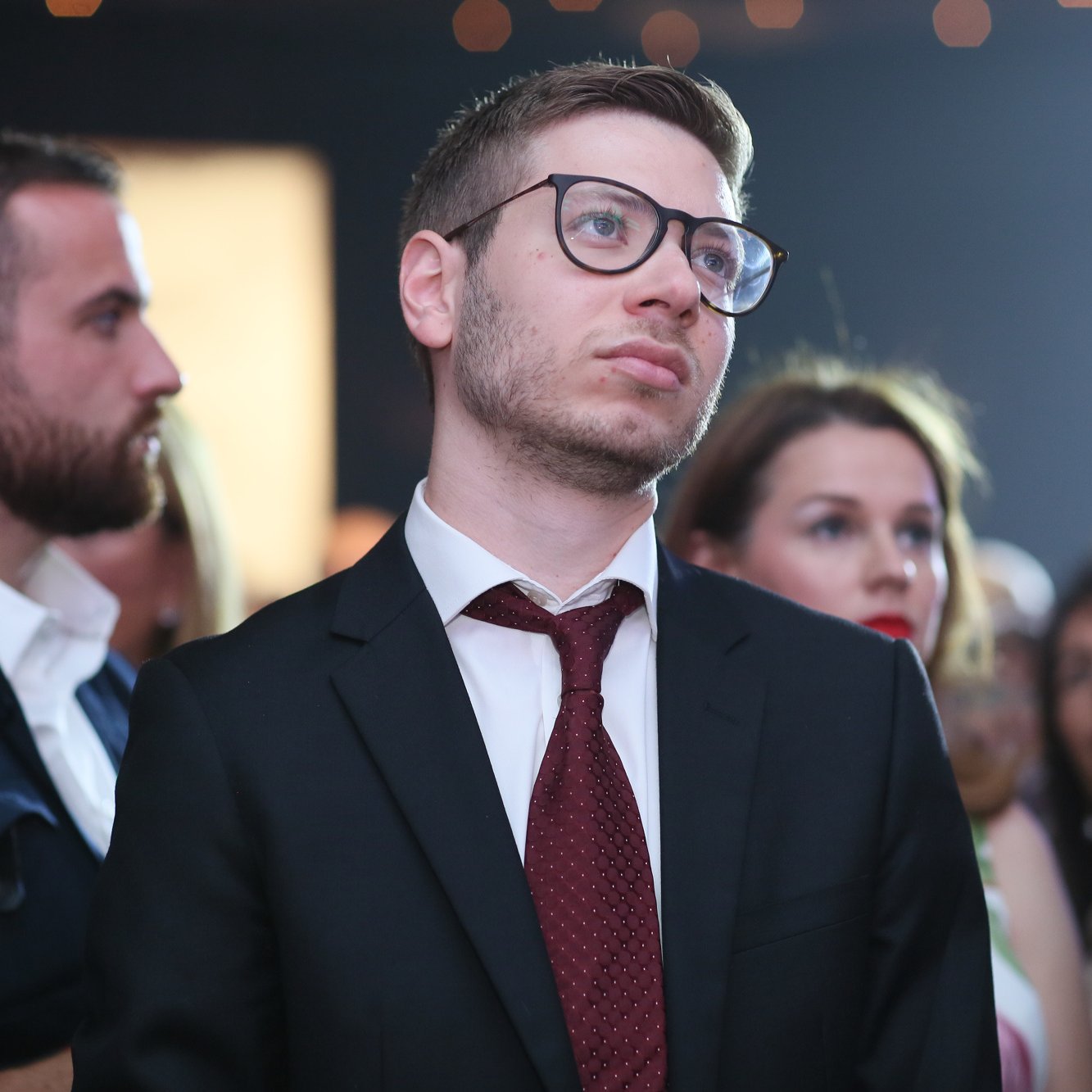 L'image montre un groupe de personnes rassemblées lors d'un événement. Au premier plan, un homme porte un costume noir avec une chemise blanche et une cravate marron. Il a des lunettes et semble pensif, le regard tourné vers le haut. À l'arrière-plan, d'autres personnes, dont une femme et un homme, sont également présentes, ajoutant à l'atmosphère de l'événement. La lumière est douce, créant une ambiance élégante.