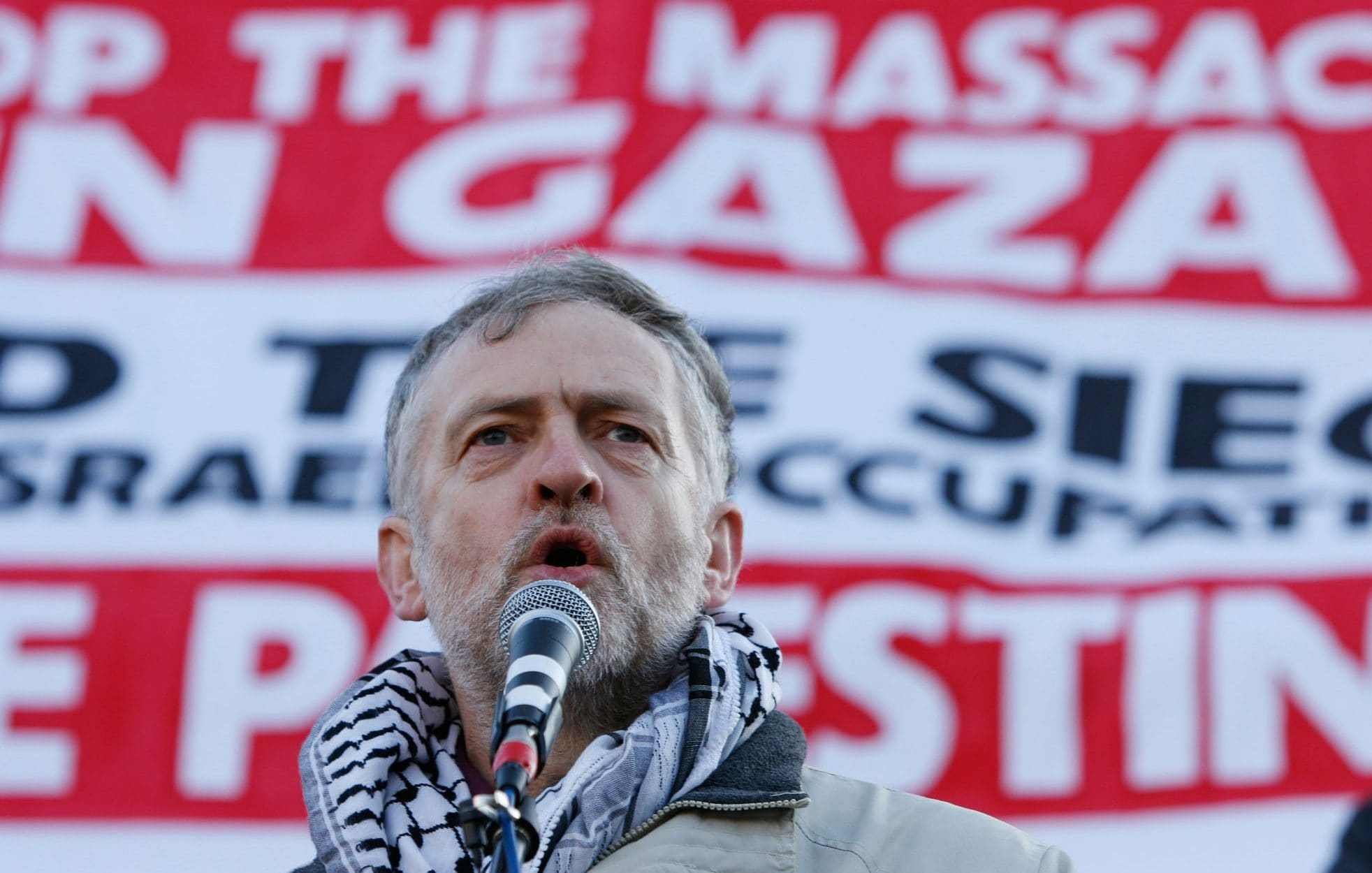 تصویر مردی را نشان می‌دهد که در حال سخنرانی است. او در مقابل یک بنر بزرگ با نوشته‌های واضح و بزرگ قرار دارد که بر موضوعاتی همچون جلوگیری از کشتار در غزه و حمایت از مردم فلسطین تأکید دارد. مرد عبا یا شال سنتی عربی را به دور گردن دارد و به نظر می‌رسد به شدت به موضوعی که درباره آن صحبت می‌کند، متعهد است. فضای تصویر، حالت اعتراضی و حماسی دارد.