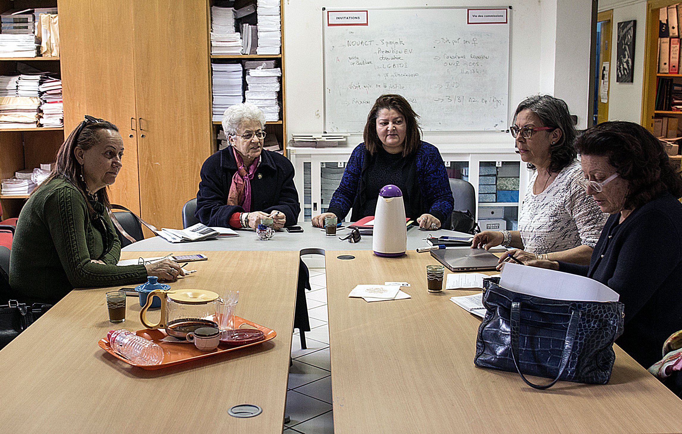 L'image montre un groupe de femmes assises autour d'une table dans une pièce qui semble être un espace de réunion. Elles sont engagées dans une discussion ou une activité collaborative. Sur la table, on peut voir des carnets, des verres et un thermos. L'environnement est simple, avec des étagères remplies de dossiers et de documents en arrière-plan. Les femmes semblent concentrées et impliquées dans leur échange.