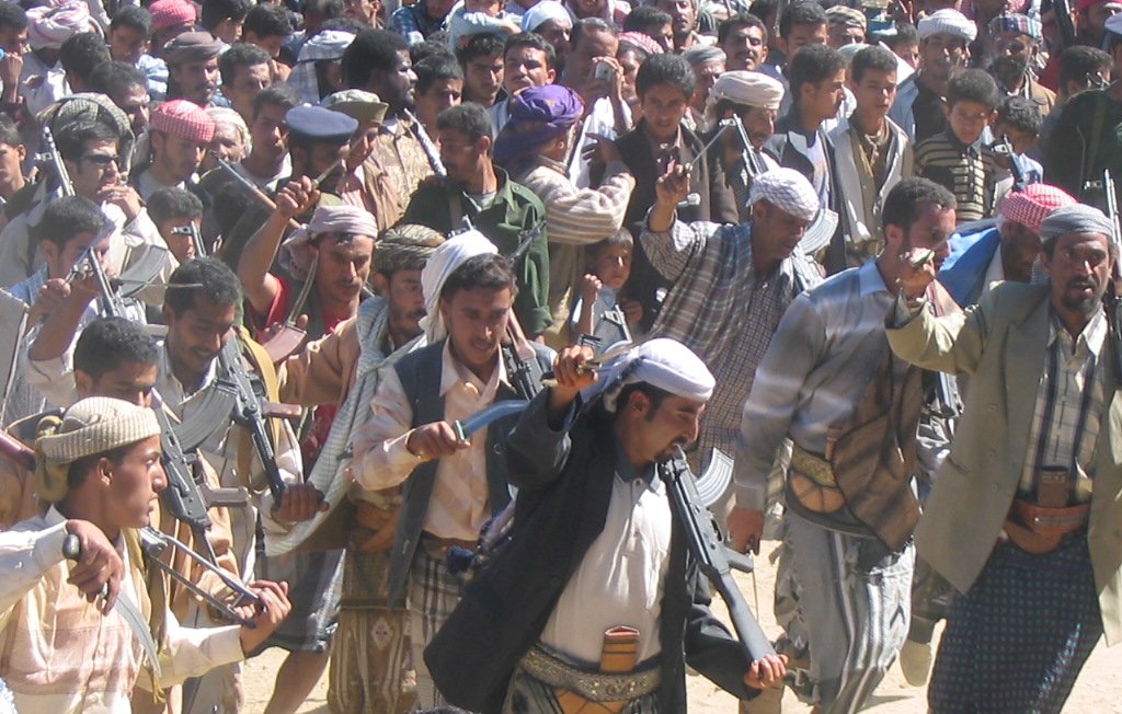L'image montre un groupe de personnes en pleine célébration ou manifestation culturelle. Les hommes portent des vêtements traditionnels, y compris des chapeaux et des écharpes. Certains sont armés avec des fusils, ce qui pourrait indiquer un contexte de fête ou de rite traditionnel dans une région où la possession d'armes est courante. L'atmosphère semble dynamique et vivante, avec les participants se déplaçant en rythme, probablement en dansant ou en chantant.